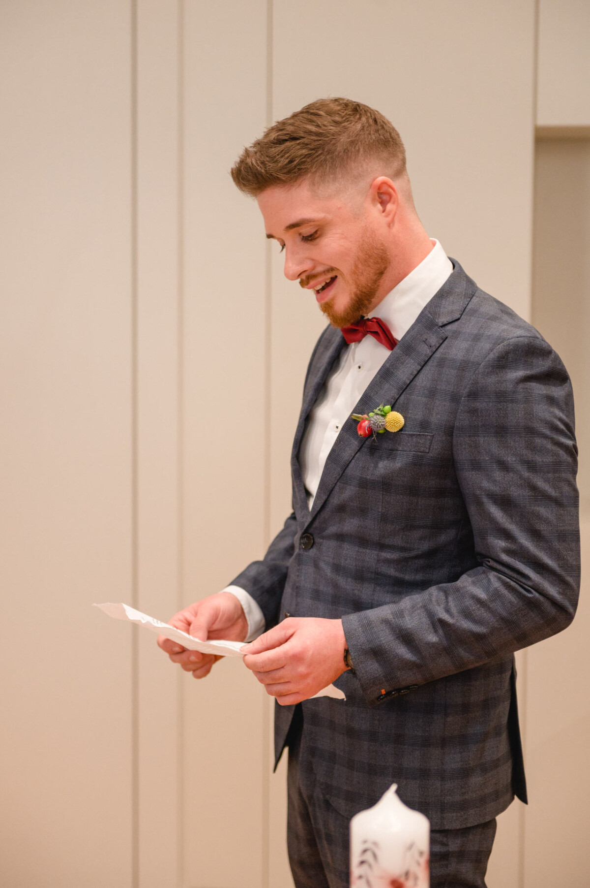 Ein Mann in einem grauen Anzug und einer roten Krawatte lächelt, während er bei einer formellen Veranstaltung eine Karte vorliest.