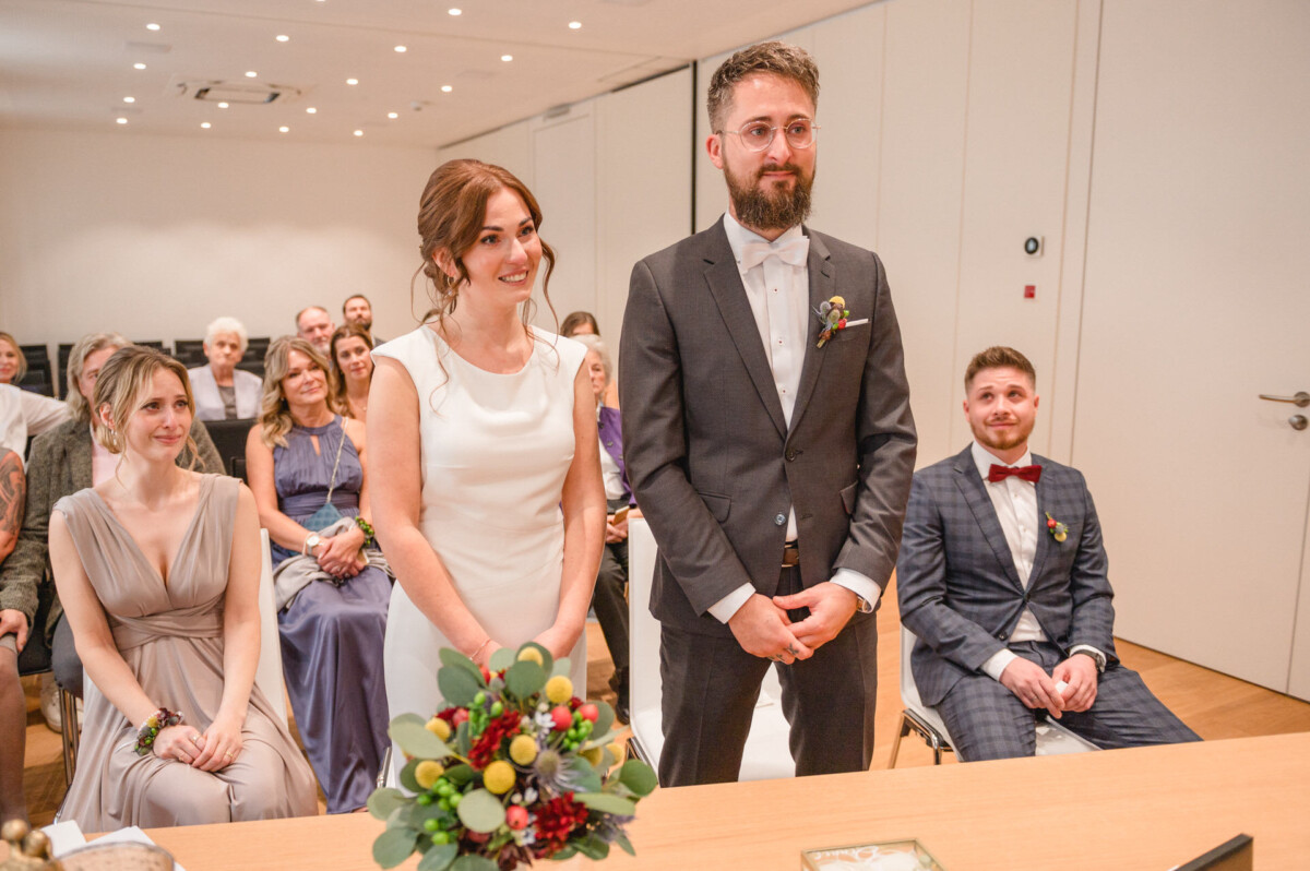 Eine Braut und ein Bräutigam lächeln bei einer Hochzeitszeremonie, im Hintergrund sitzen die Gäste.