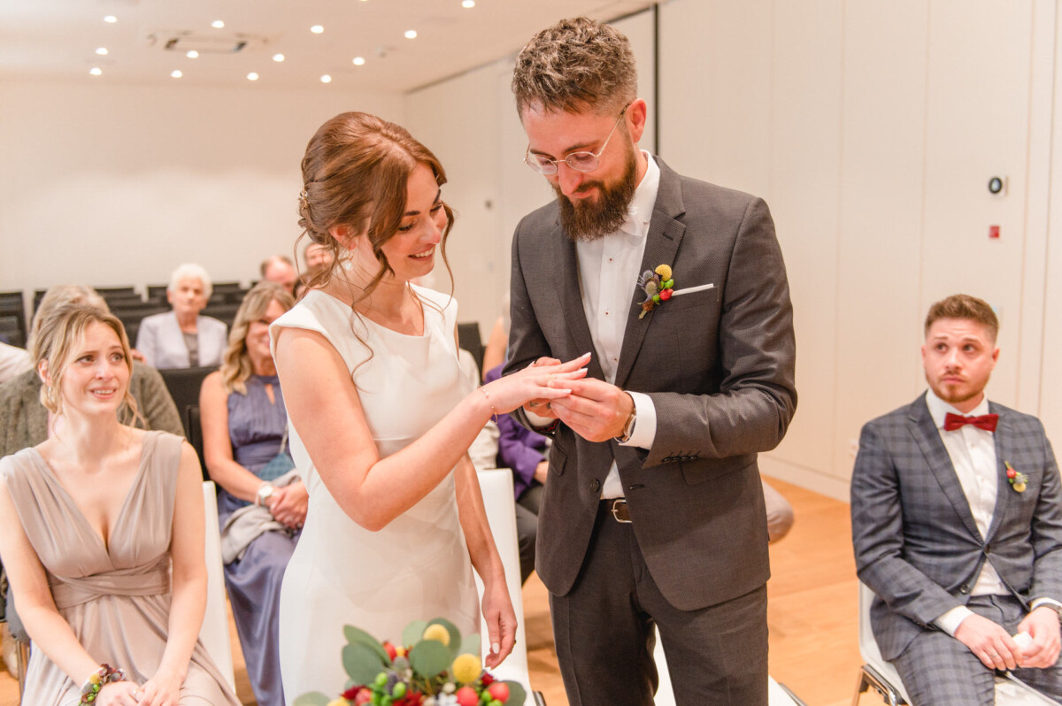 Braut und Bräutigam tauschen während einer Hochzeitszeremonie die Ringe aus, im Hintergrund schauen die Gäste zu.