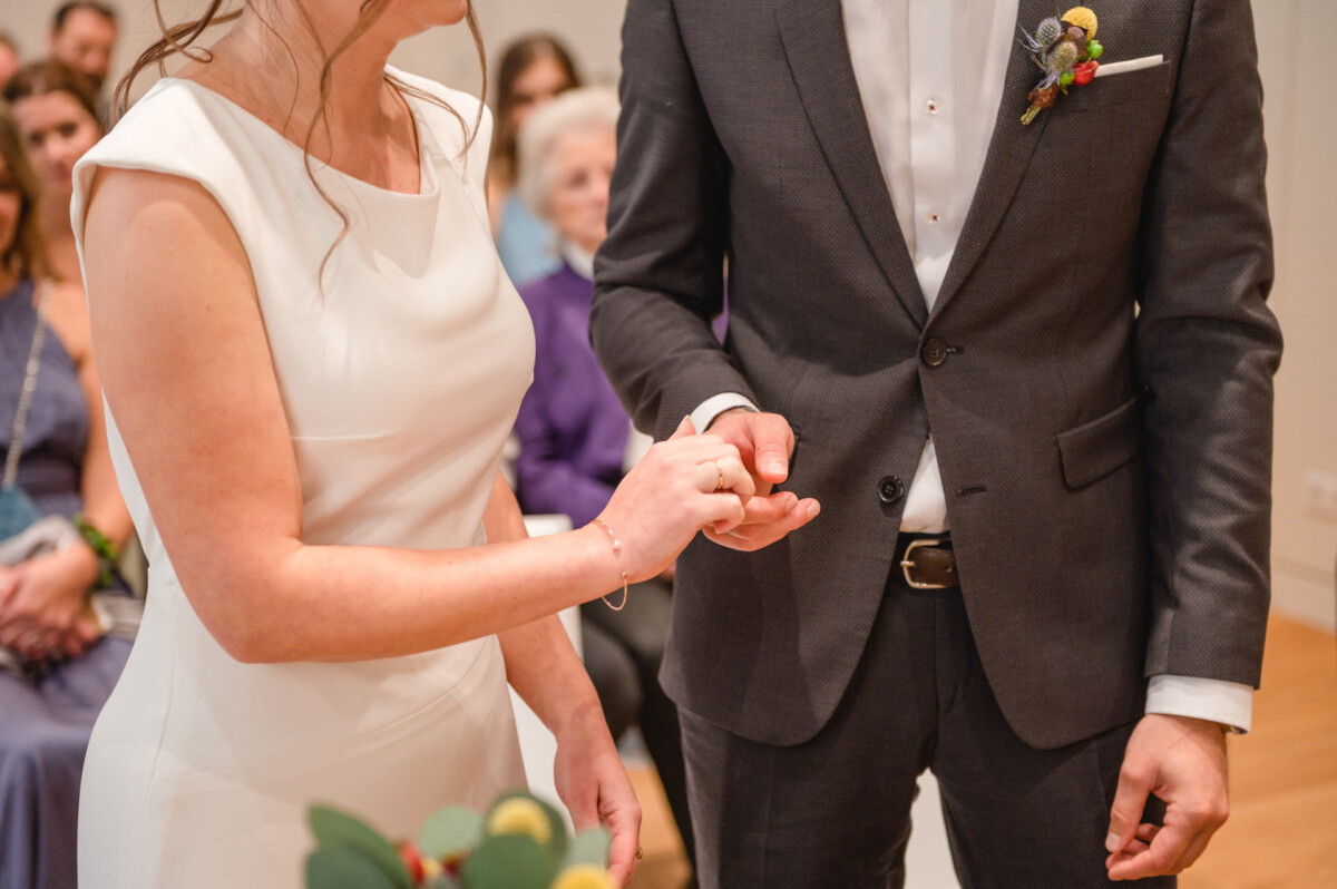 Eine Braut und ein Bräutigam halten während einer Hochzeitszeremonie mit Gästen im Hintergrund die Ringe und tauschen sie an den Händen.