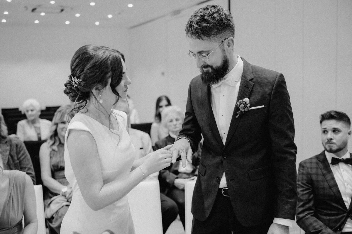 Eine Braut im weißen Kleid steckt dem Bräutigam während einer Zeremonie einen Ehering an den Finger, während die Gäste in einem gut beleuchteten Raum zuschauen.