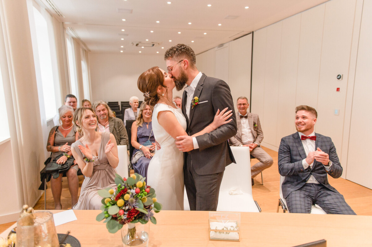 Eine Braut und ein Bräutigam küssen sich während ihrer Hochzeitszeremonie in einem Raum, während die sitzenden Gäste zuschauen und lächeln.