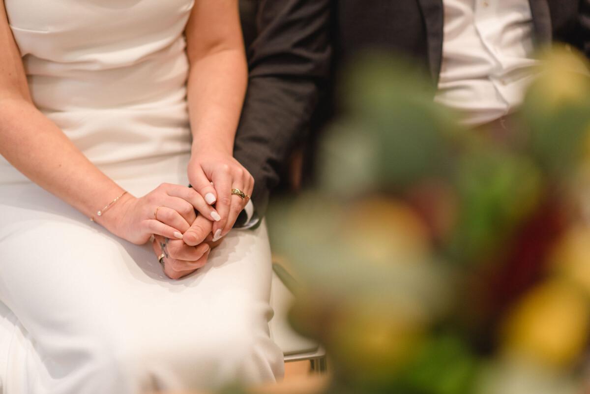Eine Nahaufnahme von Braut und Bräutigam, die im Hochzeitskleid Händchen halten, mit Fokus auf den ineinander verschlungenen Fingern und den Eheringen.