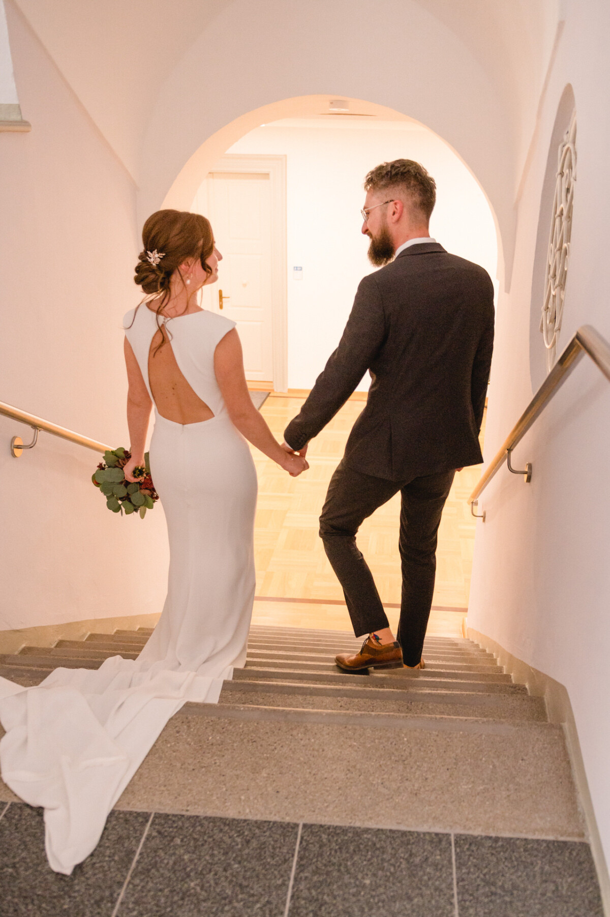 Braut und Bräutigam gehen Händchen haltend eine weiße Treppe hinunter, von hinten gesehen. Die Braut trägt ein langes weißes Kleid und der Bräutigam einen dunklen Anzug.