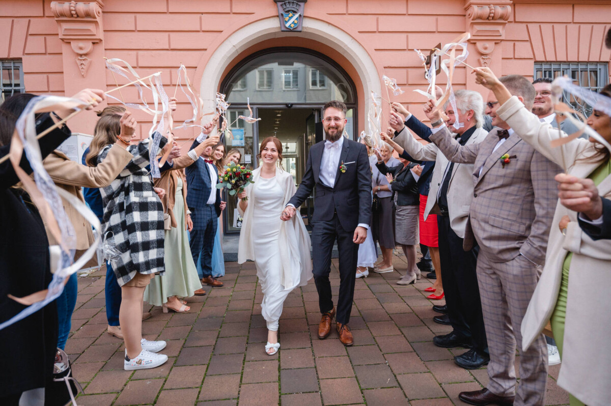 Ein frisch verheiratetes Paar geht mit Zauberstäben aus Bändern in der Hand durch eine jubelnde Menge vor einem Gebäude.