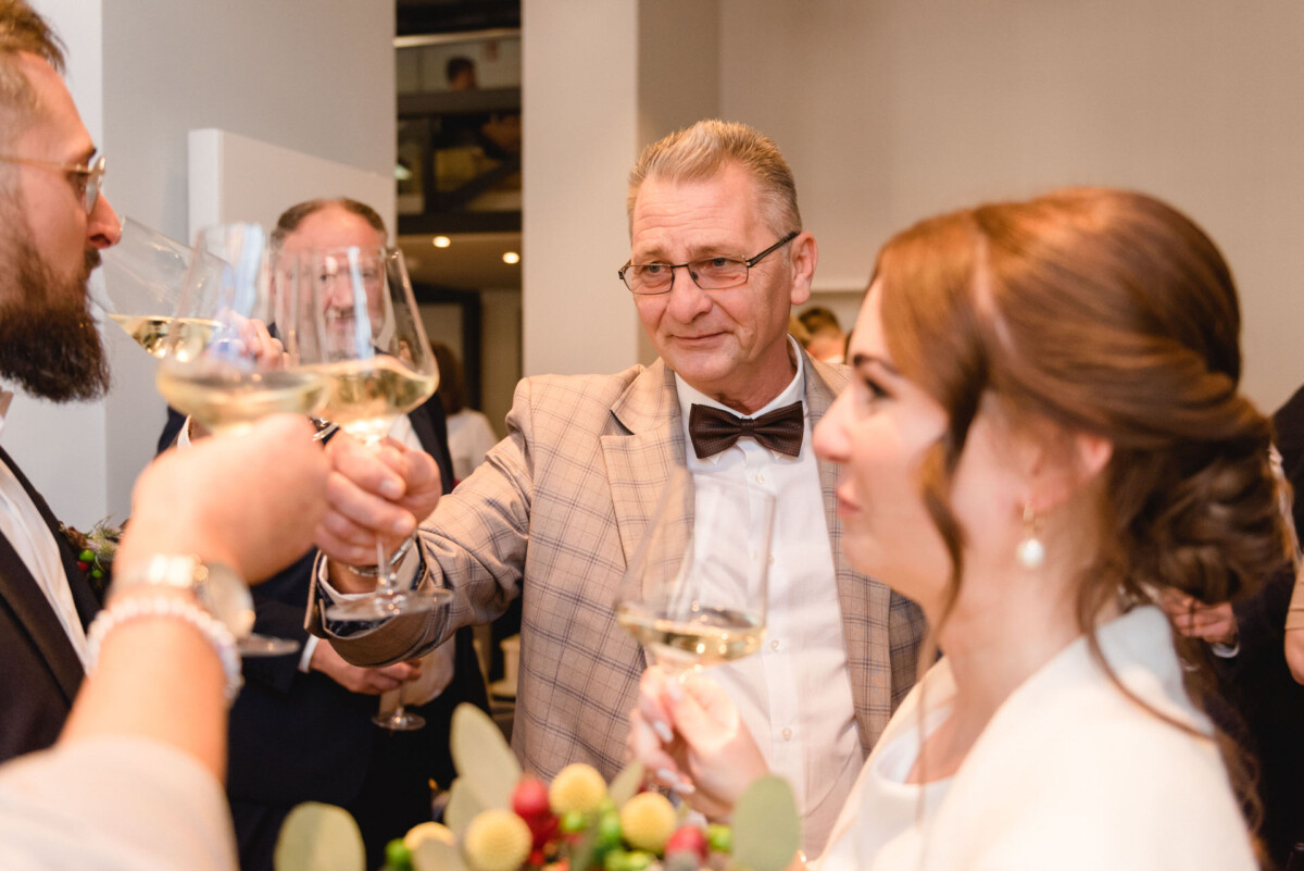 Ein älterer Mann in kariertem Anzug und Fliege stößt bei einer gesellschaftlichen Veranstaltung mit einem jüngeren Paar mit Weißweingläsern an.