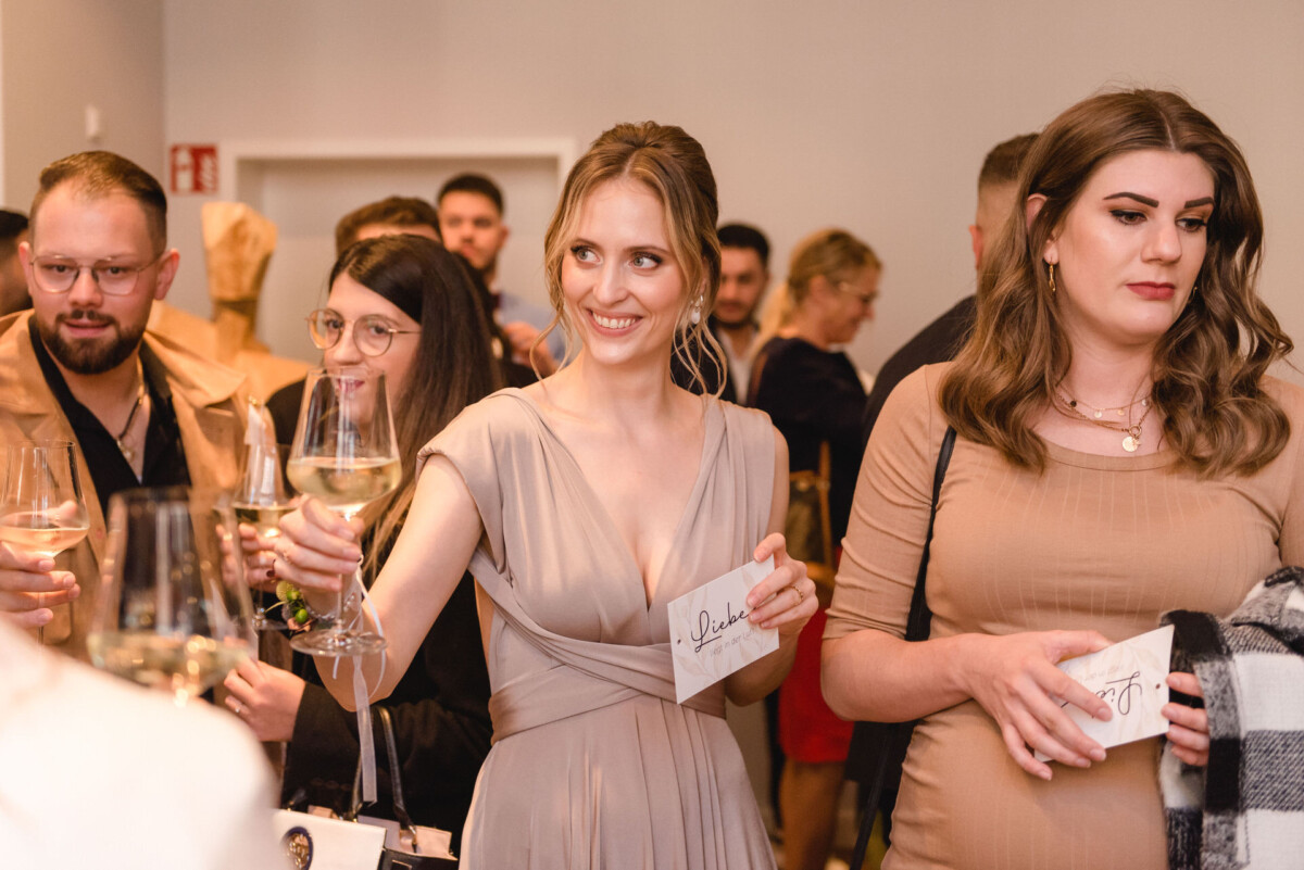 Eine Gruppe Erwachsener bei einer gesellschaftlichen Veranstaltung, die Gläser Wein in den Händen hält und miteinander plaudert, im Mittelpunkt steht eine Frau in einem hellgrauen Kleid, die in die Kamera lächelt.