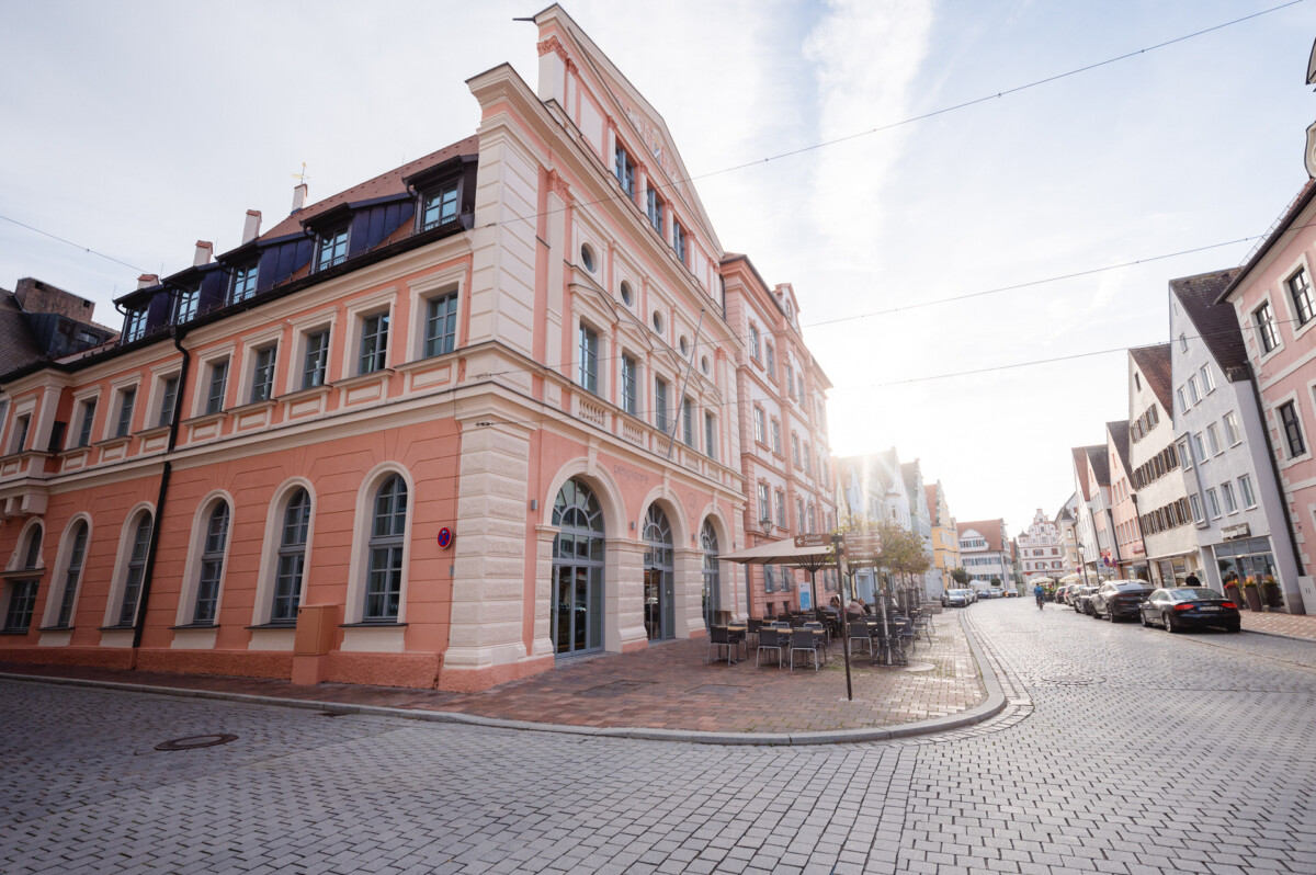 Ein sonniger europäischer Stadtplatz mit einem klassischen Gebäude in Rosa und Beige mit Bogenfenstern, einer Kopfsteinpflasterstraße, geparkten Autos und einem Fernblick auf eher traditionelle Häuser.