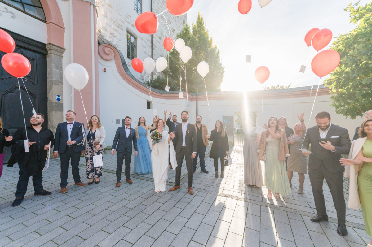 Eine Hochzeitsgruppe lässt vor einer Kirche freudig Luftballons steigen, während die Sonne in die Szene strömt.