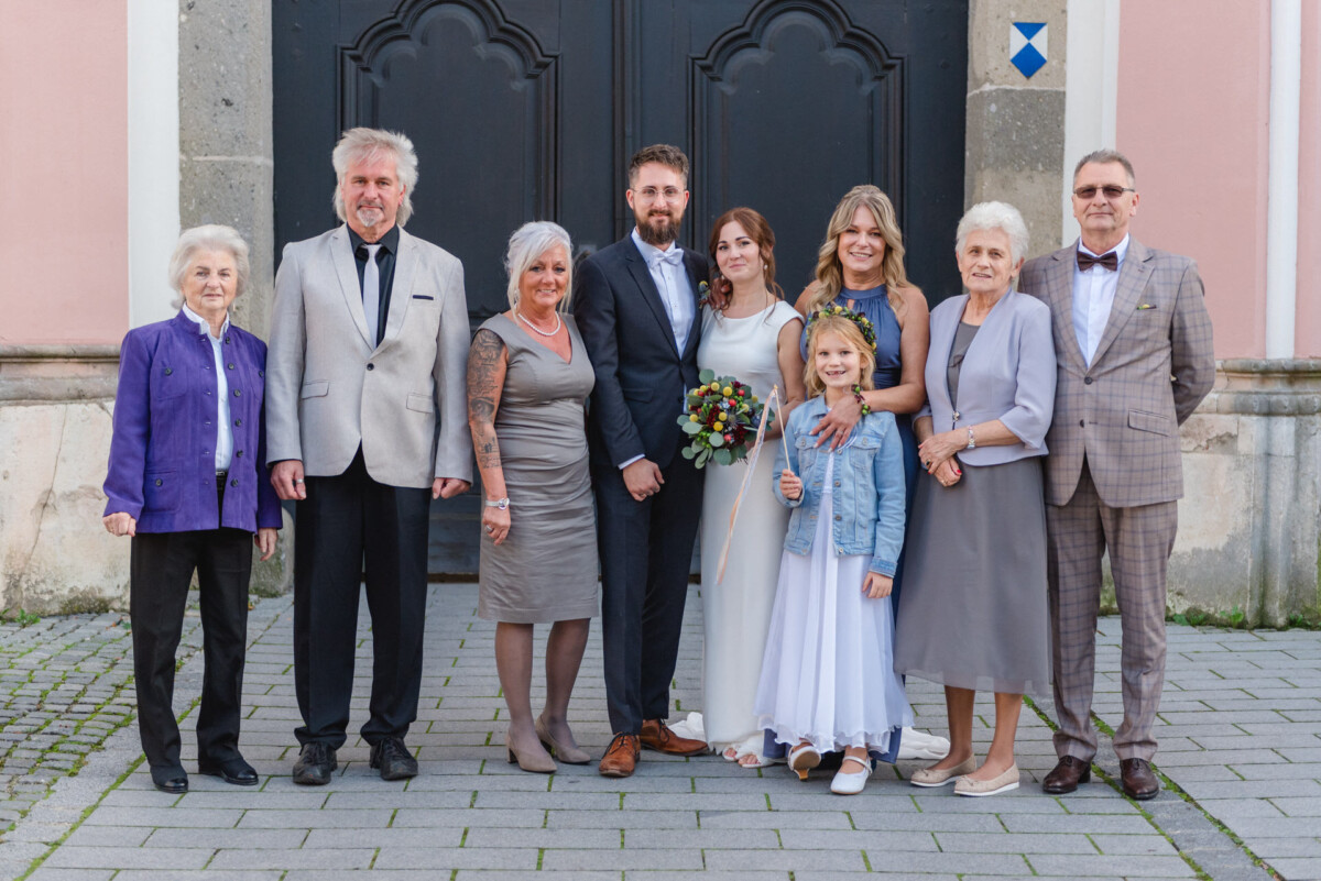 Eine neunköpfige Familie, darunter ein Kind, in Abendgarderobe, posiert gemeinsam vor dem Eingang einer Kirche.