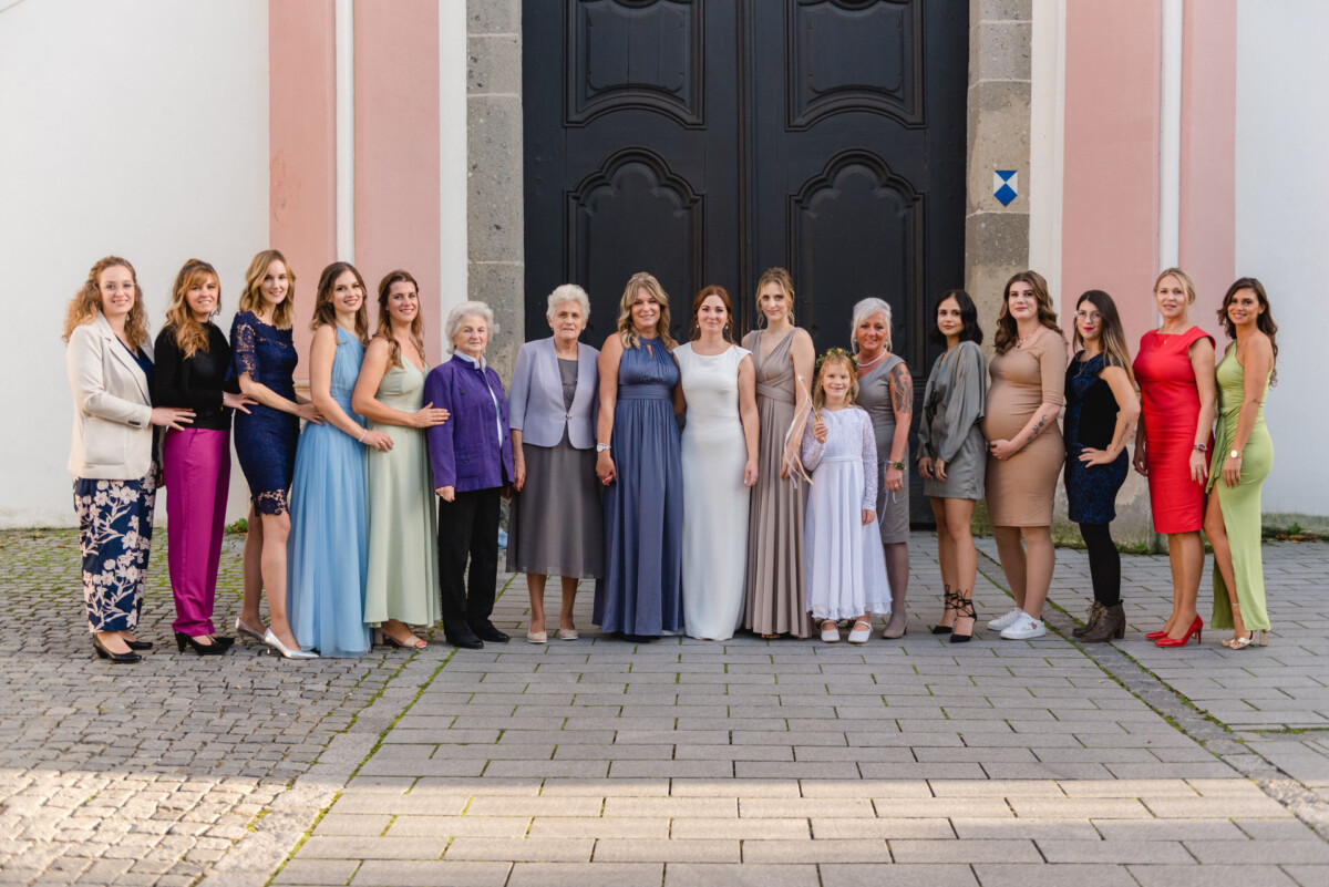Eine Gruppe von fünfzehn Frauen unterschiedlichen Alters, in Abendgarderobe gekleidet, stehen zusammen und lächeln vor einer großen Kirchentür.
