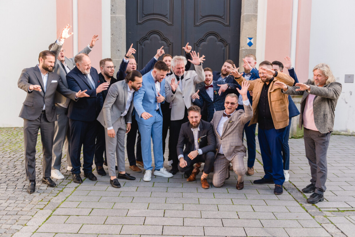 Eine Gruppe von Männern in Anzügen feiert freudig vor dem Eingang einer Kirche, einige zeigen mit dem Finger auf die Menschen und jubeln.