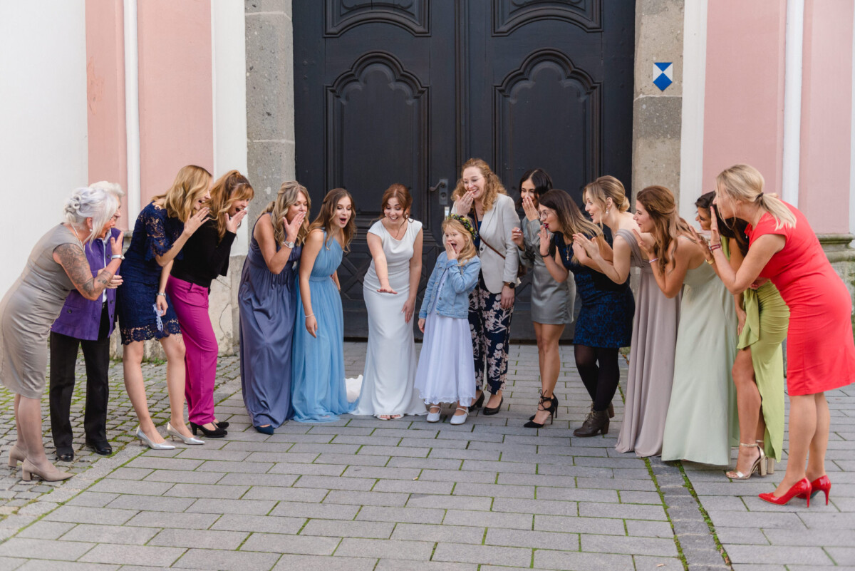 Eine Gruppe von Frauen in Abendkleidung jubelt einem jungen Mädchen in der Mitte zu und lächelt es an. Das Mädchen steht vor einem Gebäude mit Doppeltüren.