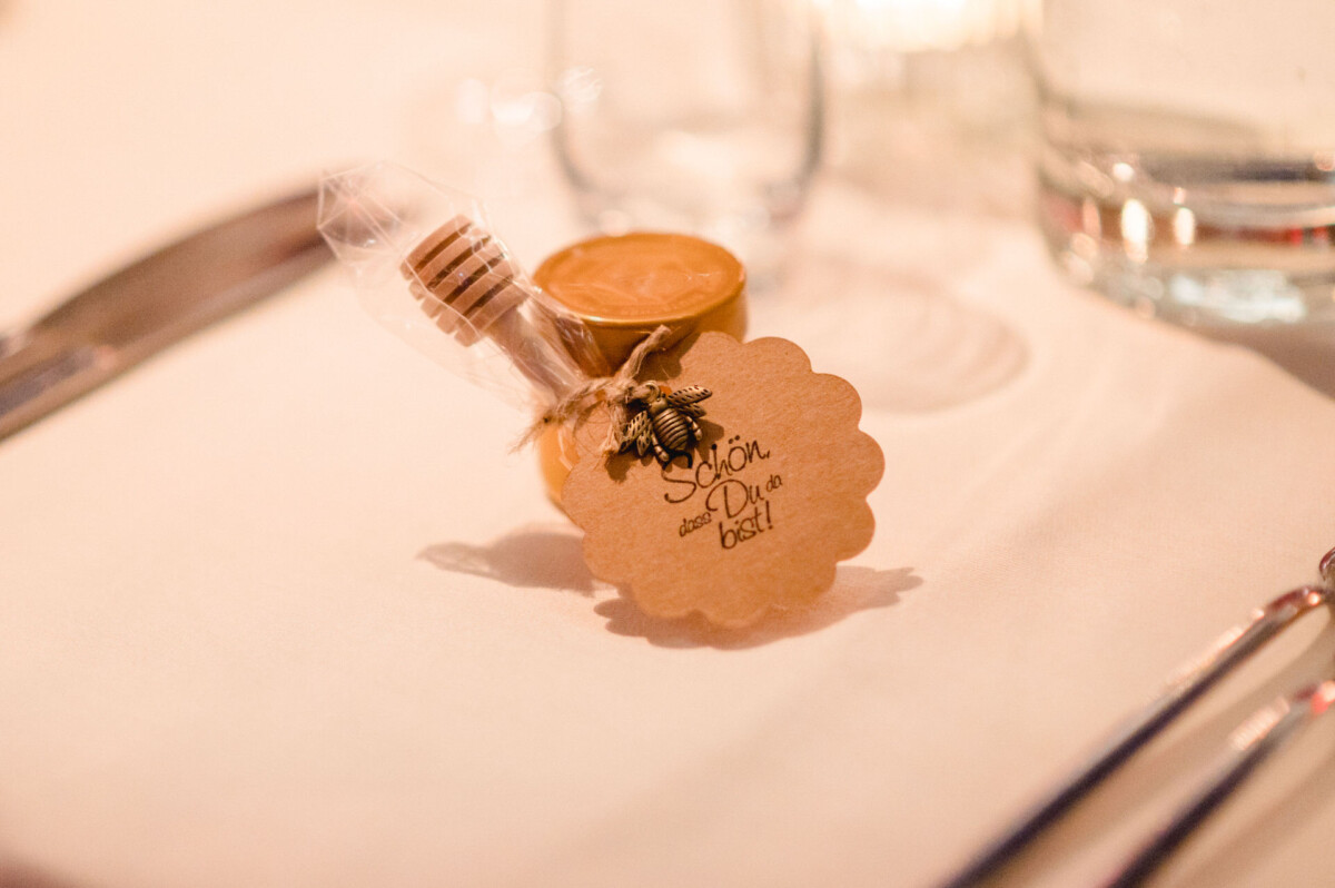 Ein Hochzeitsgeschenk bestehend aus einer kleinen Flasche Honig mit einem Bienenanhänger auf einem Schild mit der Aufschrift „Meant to Bee“ auf einem Esstisch.