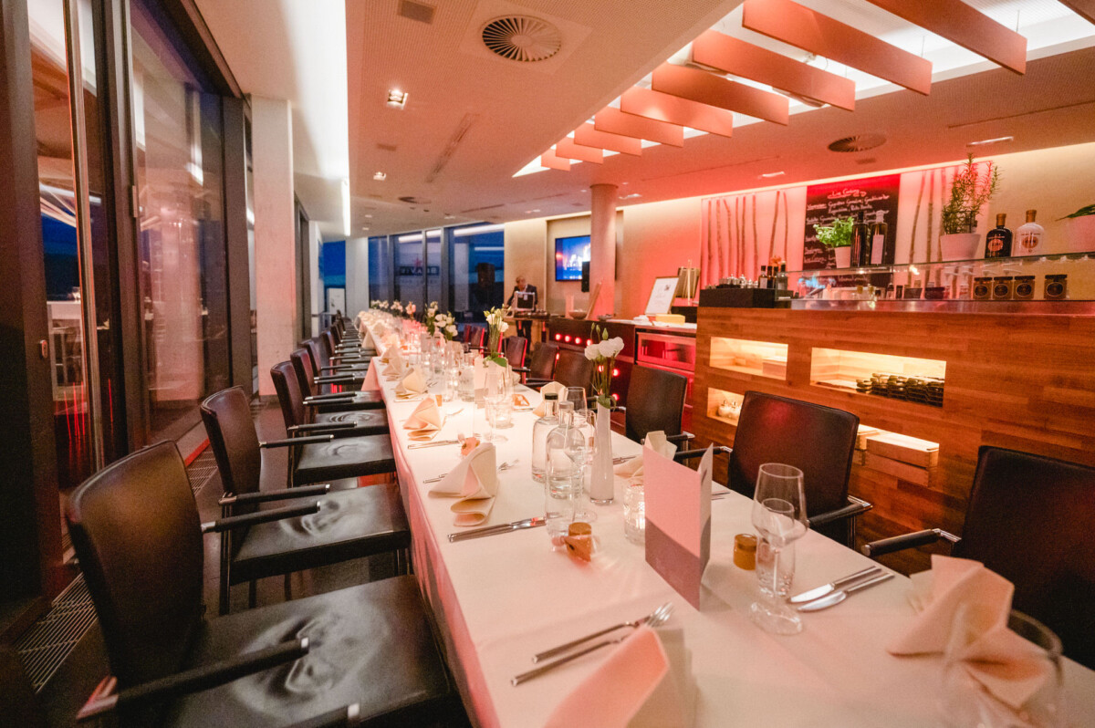 Innenansicht eines modernen Restaurants mit einem langen, für eine Mahlzeit gedeckten Esstisch, stimmungsvoller Beleuchtung und einer Bar im Hintergrund.