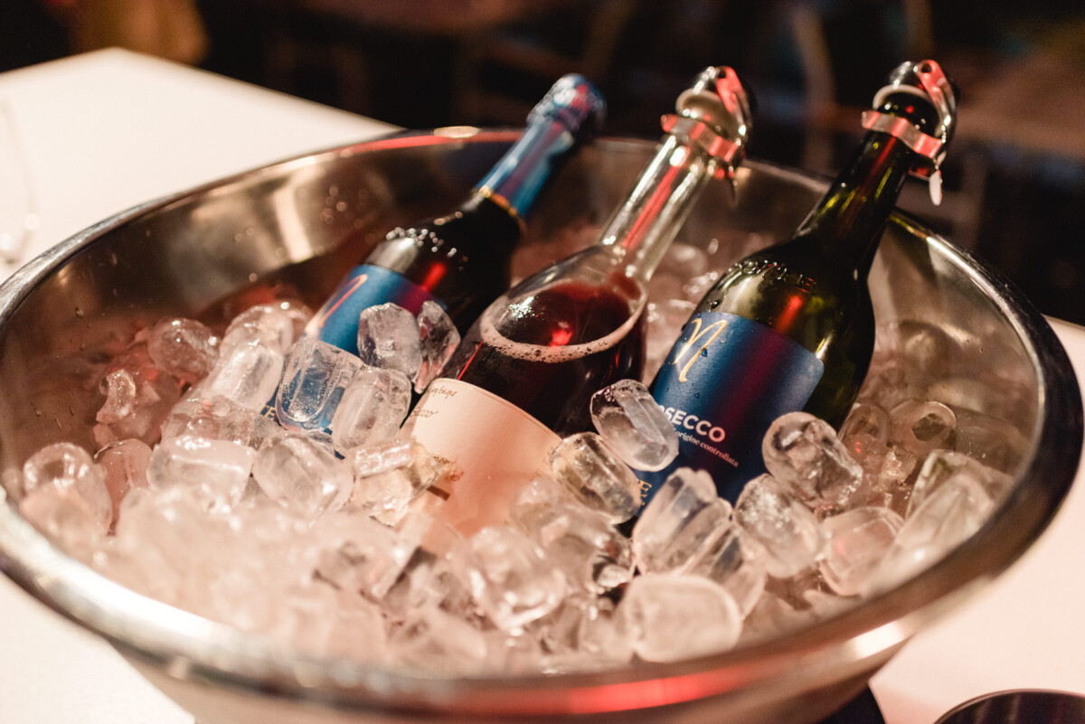 Drei Flaschen in einem mit Eis gefüllten Eimer auf einem Tisch, davon eine Flasche Rotwein und zwei Flaschen Sekt, sichtbar gekühlt.