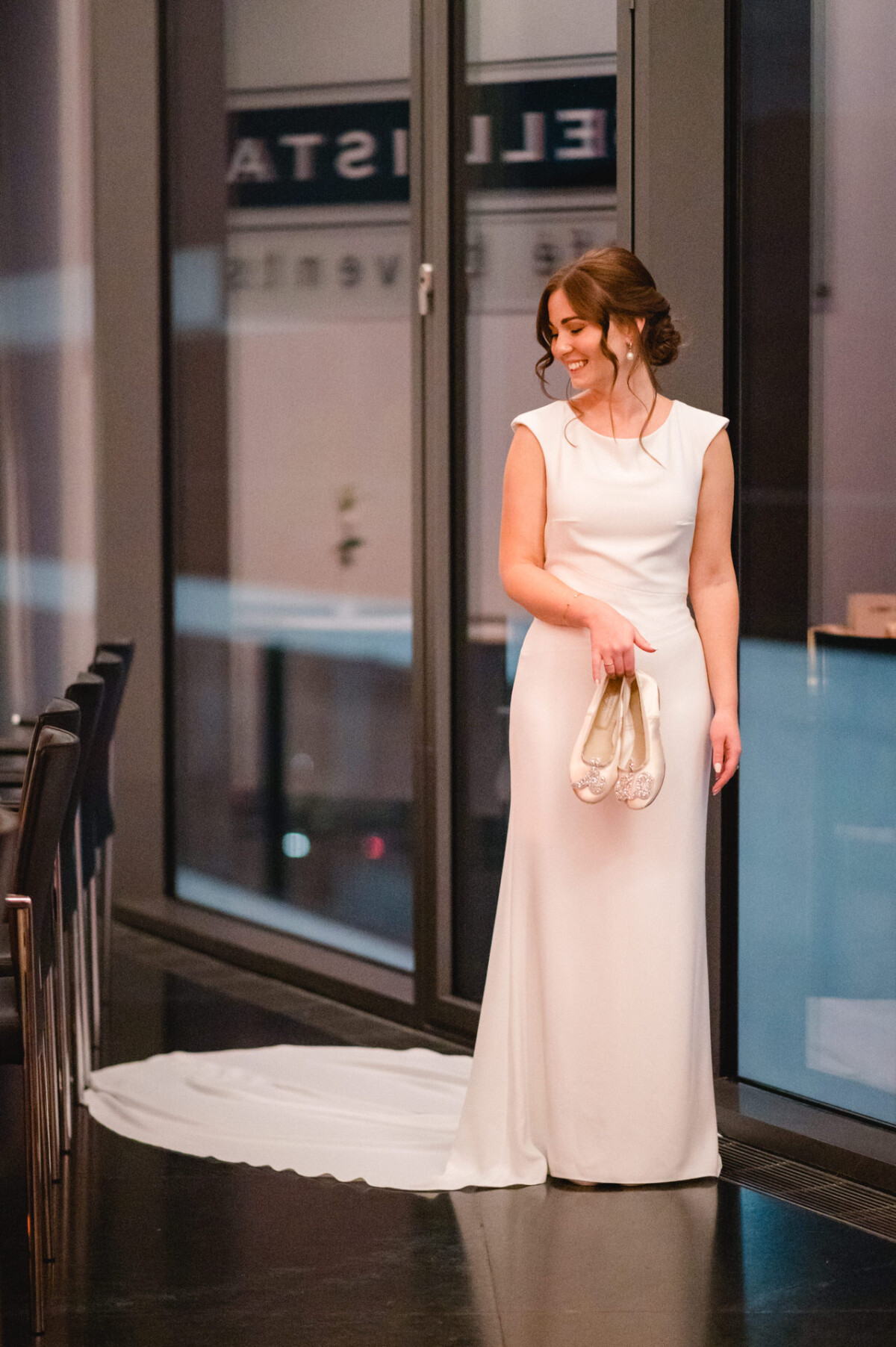 Eine Braut in einem weißen Kleid lächelt und hält ihre Schuhe. Sie steht im Innenbereich mit Glastüren im Hintergrund.