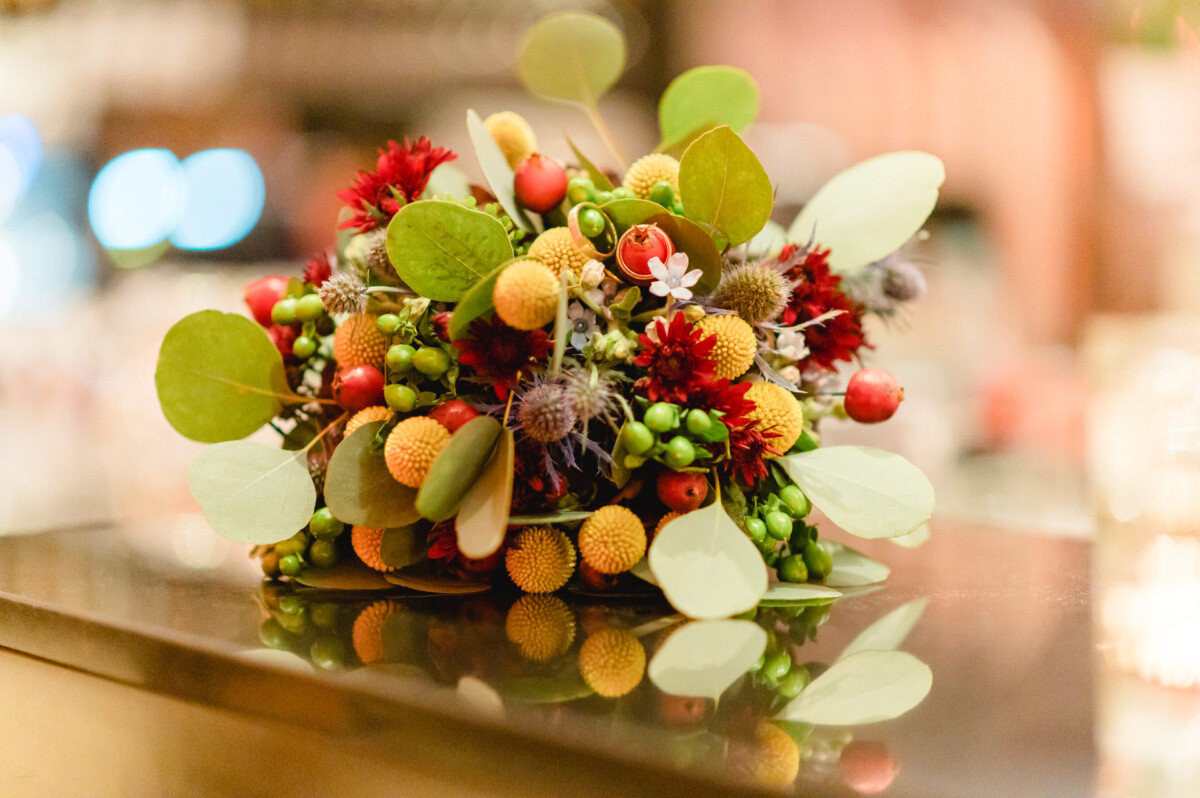 Ein farbenfroher Blumenschmuck mit einer Auswahl an roten Beeren, grünen Blättern und orangefarbenen und gelben Miniaturblumen, präsentiert auf einer reflektierenden Oberfläche.