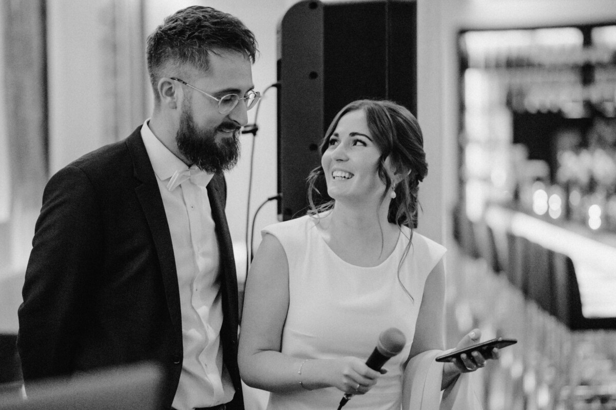 Ein monochromes Bild eines bärtigen Mannes im Anzug und einer lächelnden Frau im weißen Kleid, die ein Mikrofon hält und einen freudigen Moment teilt.