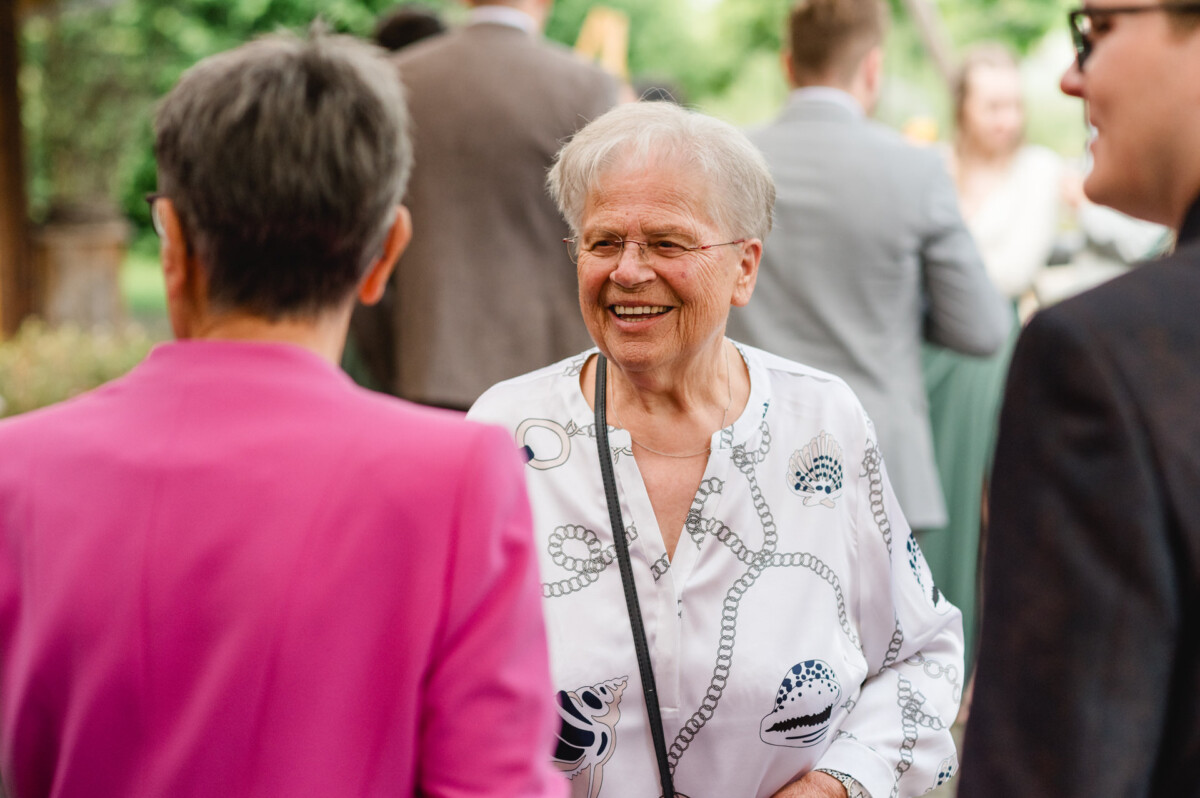 2024.10.05-Lea-Viktor-Hochzeit-Schammenhof-Langenau-bei-Ulm-00686 Eine ältere Person in einem gemusterten Hemd lächelt herzlich, während sie sich bei einem Treffen im Freien in der Nähe des malerischen Schammenhofs, etwas außerhalb von Langenau und Ulm, mit anderen unterhält.