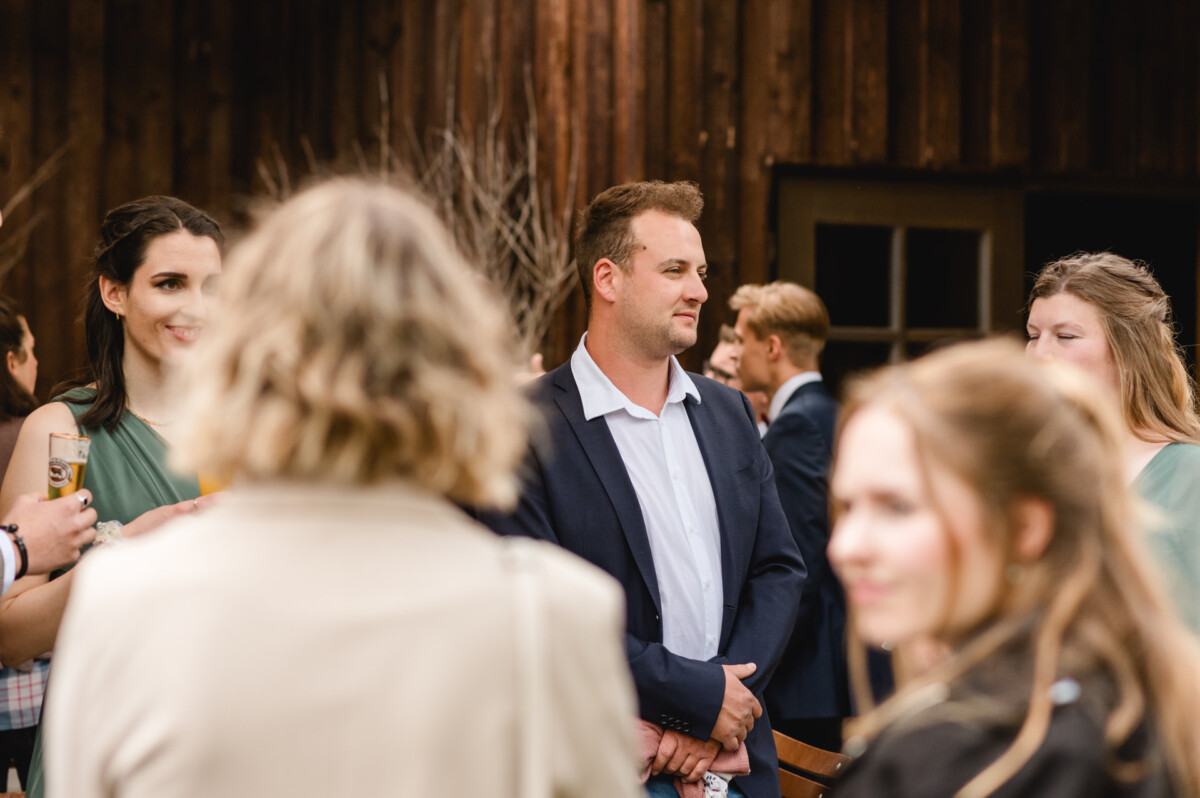 2024.10.05-Lea-Viktor-Hochzeit-Schammenhof-Langenau-bei-Ulm-00689 Bei einer zauberhaften Open-Air-Veranstaltung auf dem Schammenhof in Langenau steht ein Mann im Anzug im Mittelpunkt der Gesellschaft. Das rustikale Holzgebäude dient als charmante Kulisse und bietet den perfekten Rahmen für diese unvergessliche Hochzeitsfeier in der Nähe von Ulm.