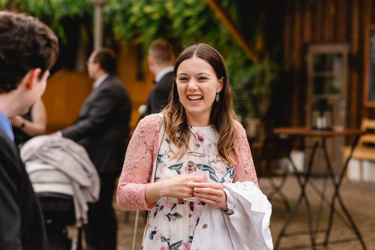 Bei einer Hochzeit im Schammenhof hält eine Frau in einem geblümten Kleid mit rosa Spitzenärmeln ein Getränk und einen weißen Mantel in der Hand und lächelt inmitten von Menschen und Grün in Langenau bei Ulm. Bei einer Hochzeit im Schammenhof hält eine Frau in einem geblümten Kleid mit rosa Spitzenärmeln ein Getränk und einen weißen Mantel in der Hand und lächelt inmitten von Menschen und Grün in Langenau bei Ulm.