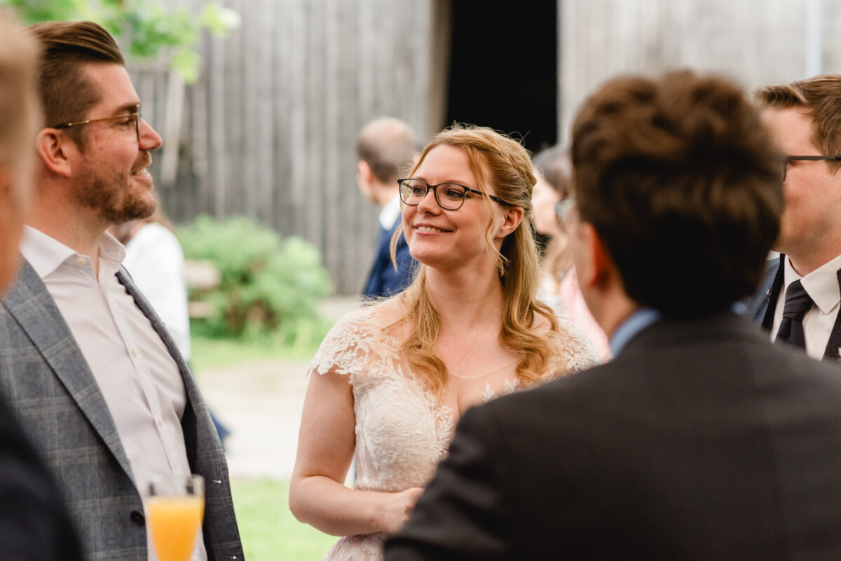 Bei der Hochzeit im Schammenhof lächelt eine Frau in einem weißen Kleid und mit Brille herzlich, während sie sich im Freien mit einer Gruppe von Menschen unterhält. Bei der Hochzeit im Schammenhof lächelt eine Frau in einem weißen Kleid und mit Brille herzlich, während sie sich im Freien mit einer Gruppe von Menschen unterhält.