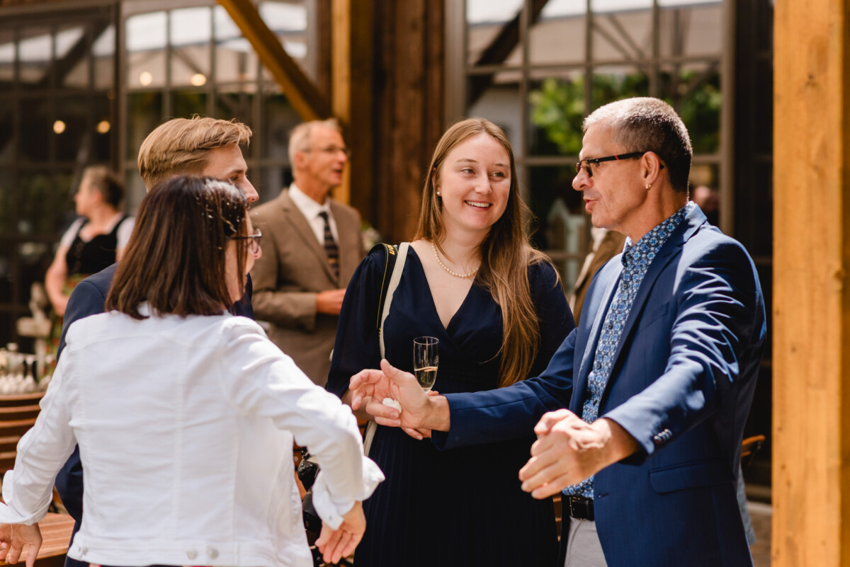 Bei einer Hochzeit im Schammenhof bei Langenau versammeln sich elegant gekleidete Gäste und unterhalten sich fröhlich im hellen Holzambiente. Eine Frau im schwarzen Kleid erhebt ihr Sektglas und trägt zur ausgelassenen Stimmung dieser unvergesslichen Ulmer Feier bei. Bei einer Hochzeit im Schammenhof bei Langenau versammeln sich elegant gekleidete Gäste und unterhalten sich fröhlich im hellen Holzambiente. Eine Frau im schwarzen Kleid erhebt ihr Sektglas und trägt zur ausgelassenen Stimmung dieser unvergesslichen Ulmer Feier bei.