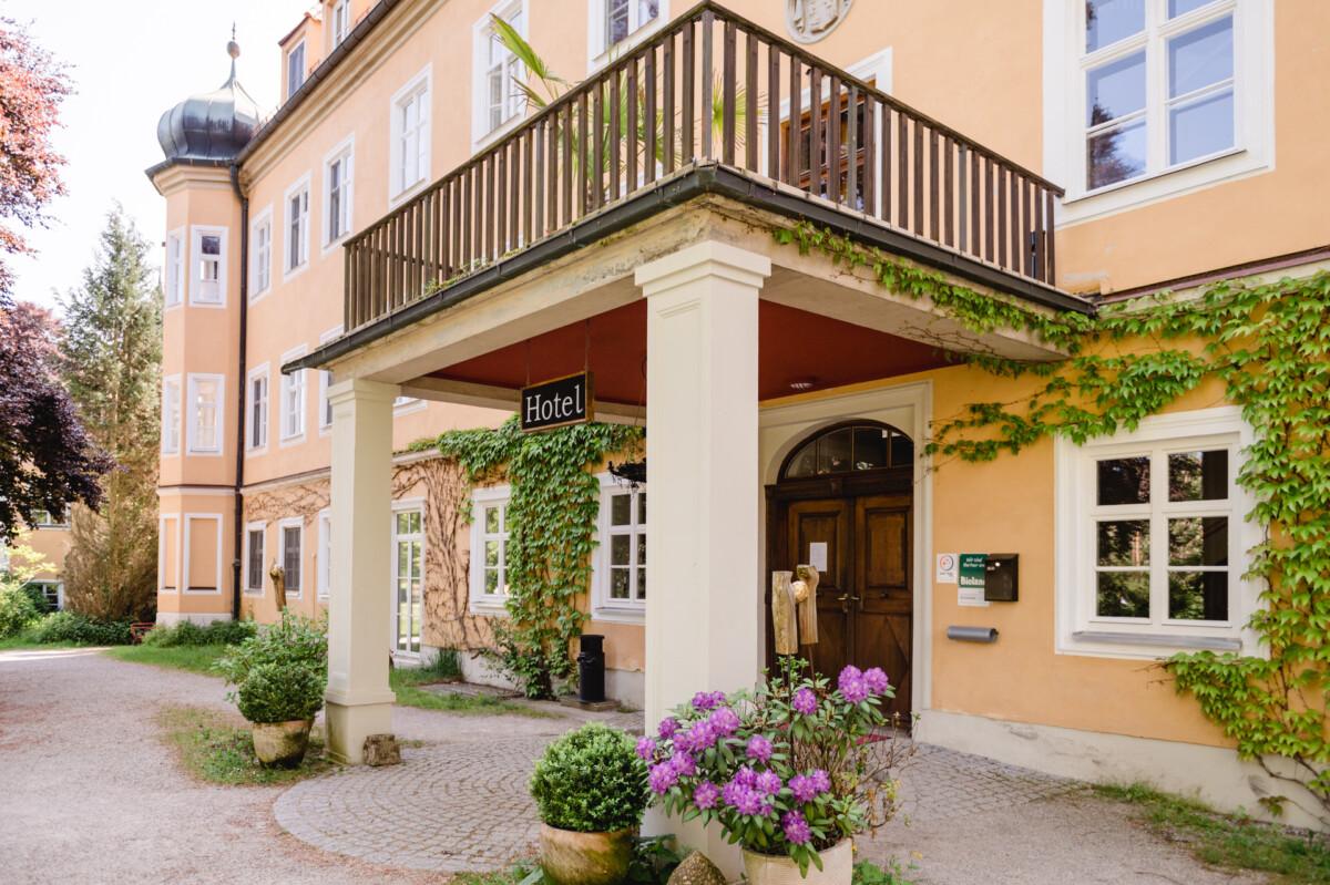 Eingang eines gelben Hotels mit Holztür und Balkon, bedeckt mit grünem Efeu. Zu sehen sind violette Topfblumen und ein „Hotel“-Schild.