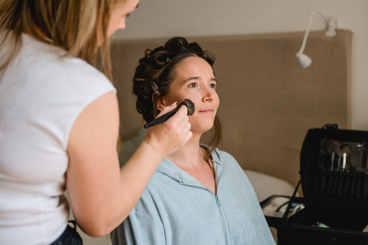 Eine Frau sitzt in einem gemütlichen Innenbereich und lässt sich von einer anderen Person mit einem Pinsel Make-up auftragen.