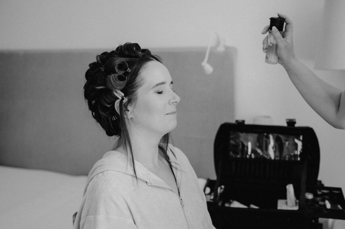 Eine Frau mit Lockenwicklern sitzt da, während jemand Haarspray aufträgt. Im Hintergrund ist ein Make-up-Set zu sehen.