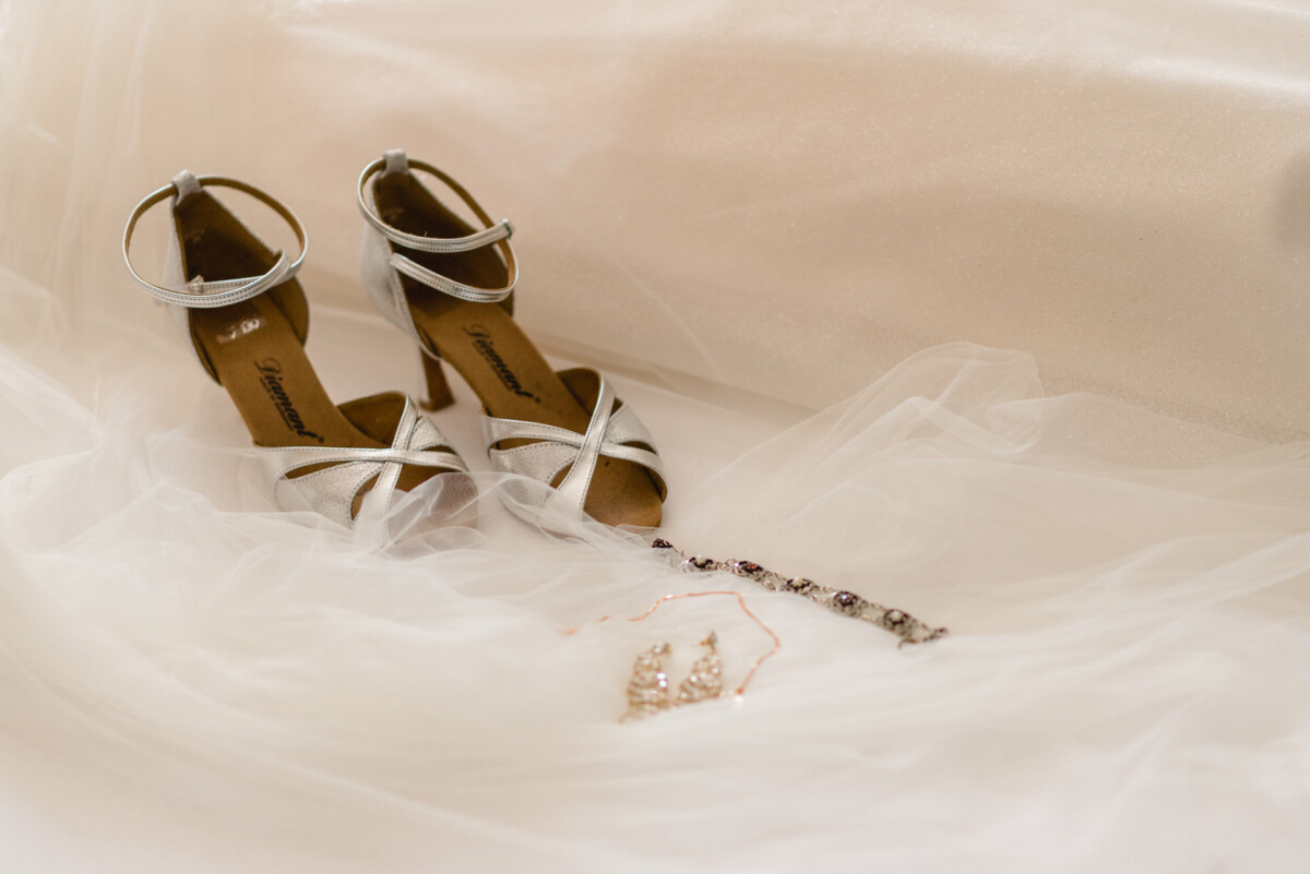 Silberne Stöckelschuhe und Schmuck, darunter eine Halskette und Ohrringe, ausgelegt auf einem weißen, durchsichtigen Stoffhintergrund.