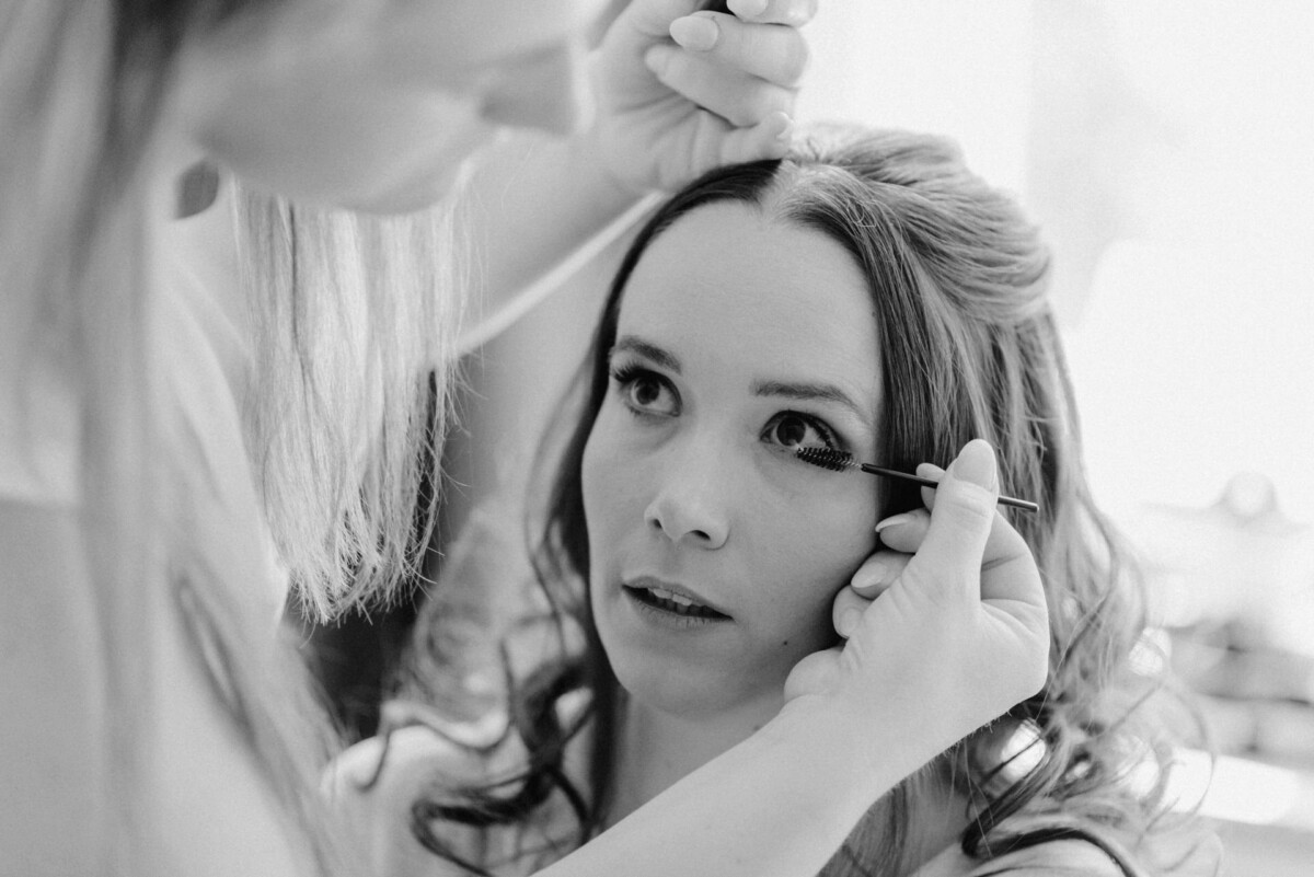 Eine Frau bekommt in einer Nahaufnahme in Schwarzweiß von einer anderen Person Mascara auf die Wimpern aufgetragen.