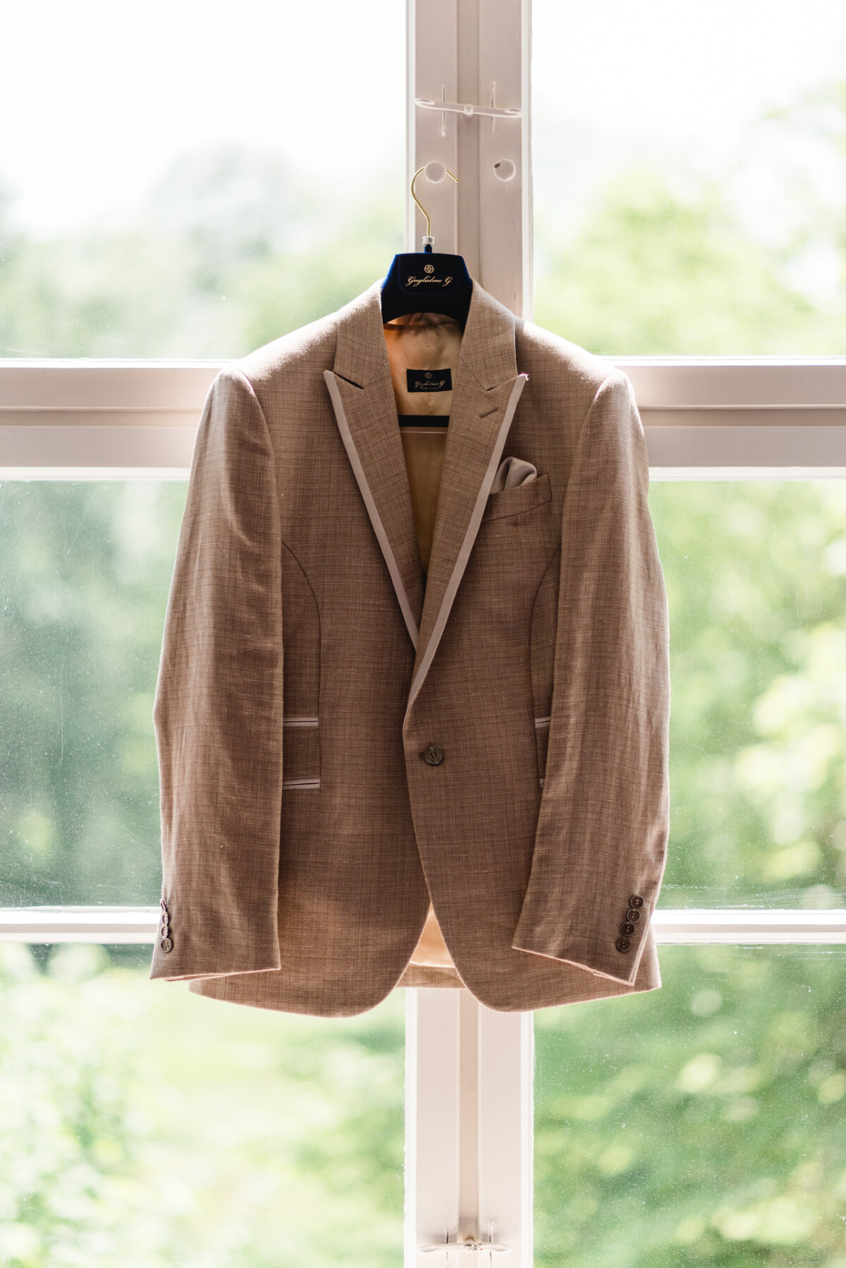 Ein beiges Jackett hängt auf einem Kleiderbügel vor einem Fenster. Draußen sieht man grünes Laub.