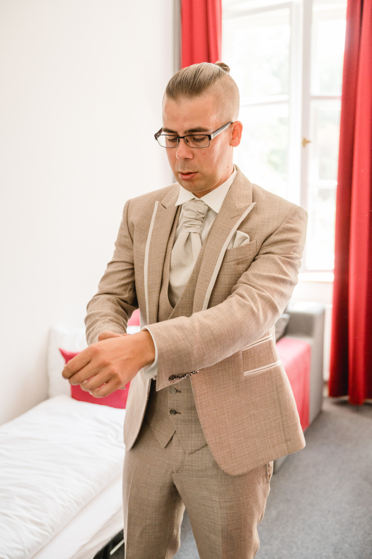 Ein Mann in beigem Anzug und Brille richtet seinen Ärmel in einem hell erleuchteten Raum mit roten Vorhängen.