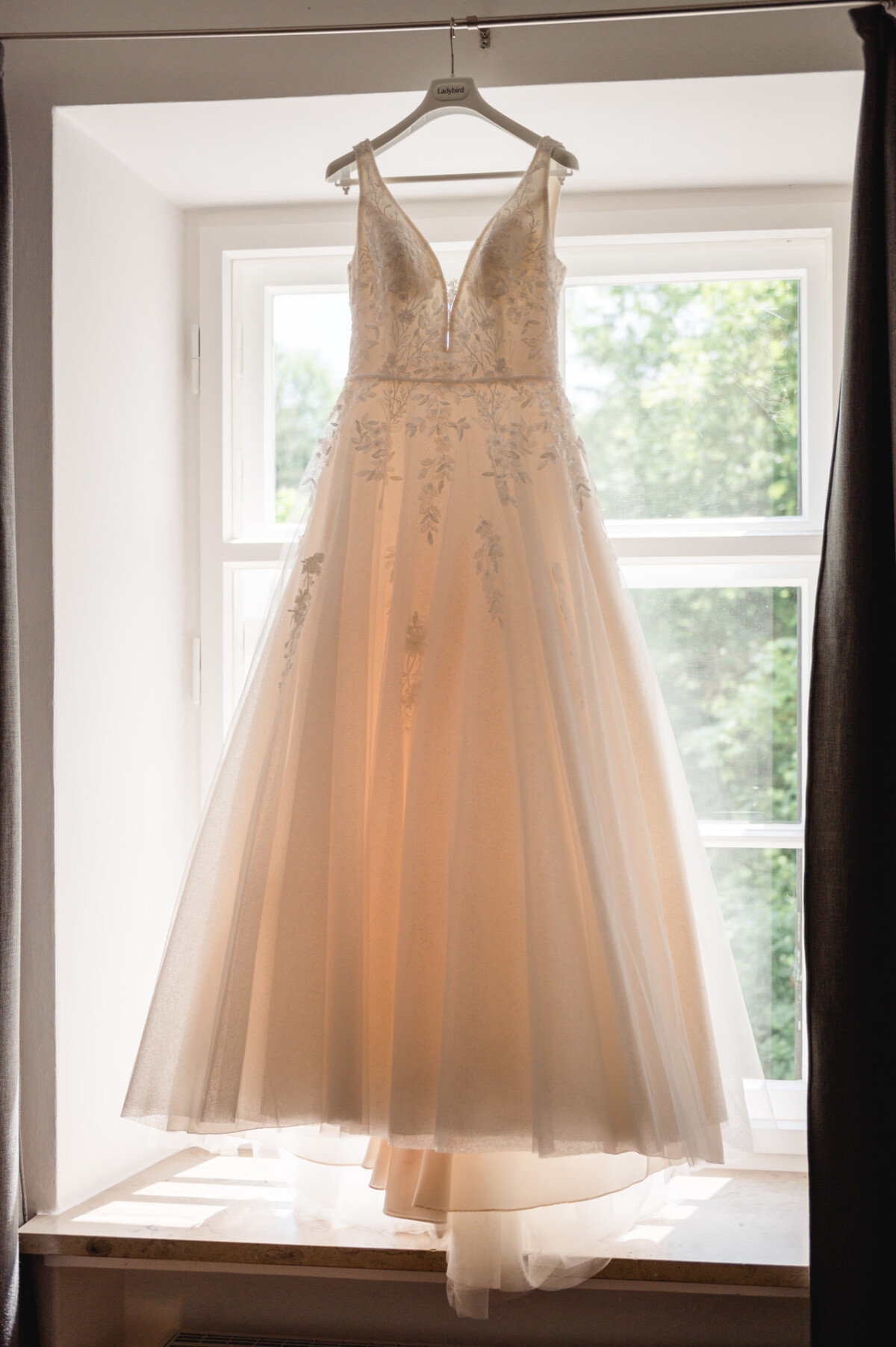 Ein weißes Brautkleid mit Blumenstickerei hängt an einem Fenster und wird vom Tageslicht erhellt.