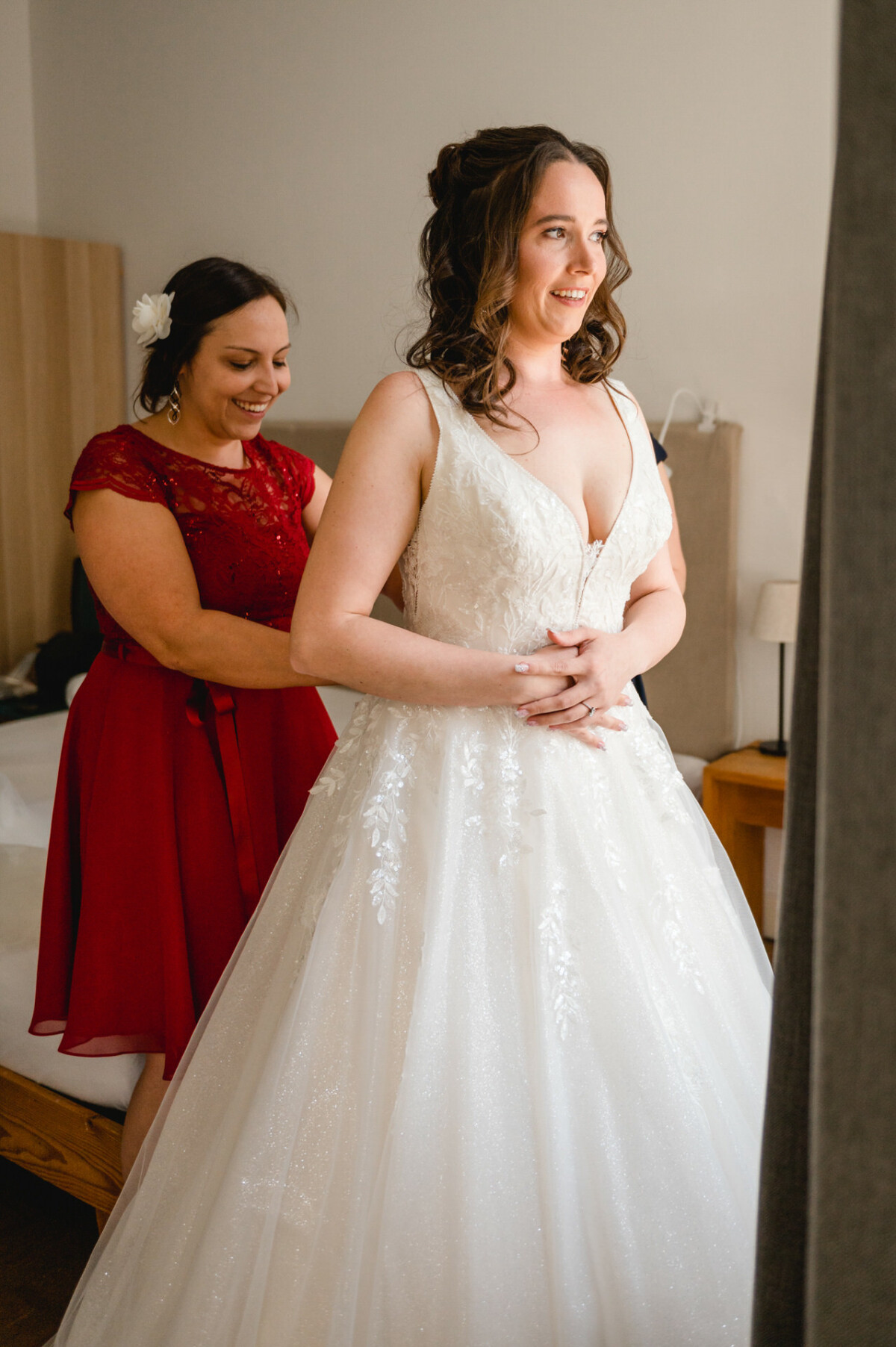 Eine Braut in einem weißen Kleid steht lächelnd neben einem Fenster. Eine Frau in einem roten Kleid hilft ihr mit dem Kleid.