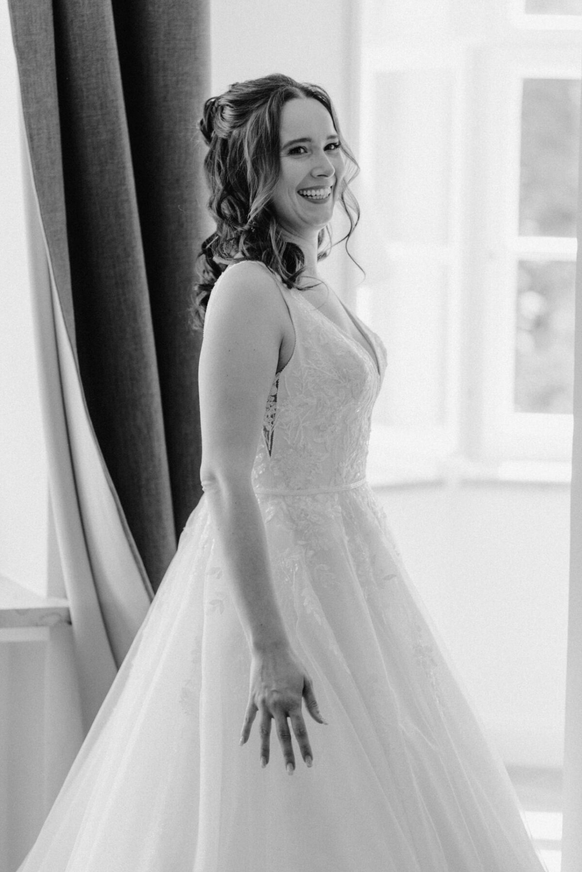Eine Frau im Brautkleid lächelt, während sie vor einem Fenster mit Vorhängen steht.