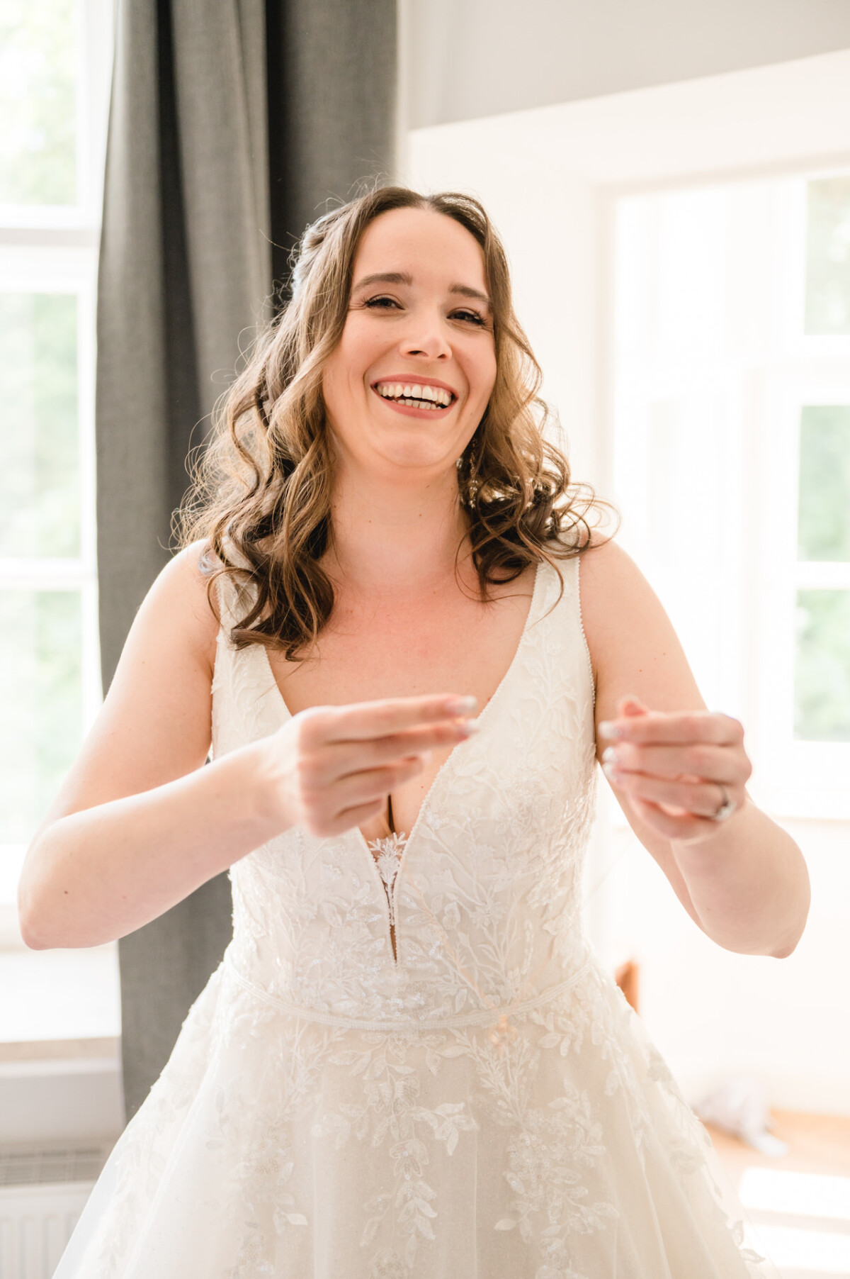 Eine Frau in einem weißen Spitzenbrautkleid lächelt, während sie im Haus neben einem Fenster mit grauen Vorhängen steht.