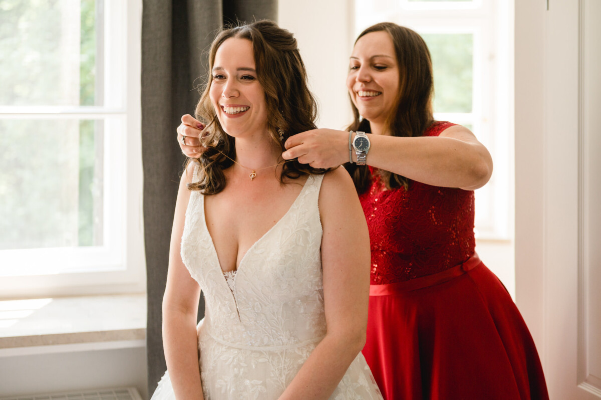 Zwei Frauen in einem gut beleuchteten Raum, eine im Brautkleid, die andere in einem roten Kleid, lächeln, während die Frau in Rot der Braut eine Halskette zurechtrückt.