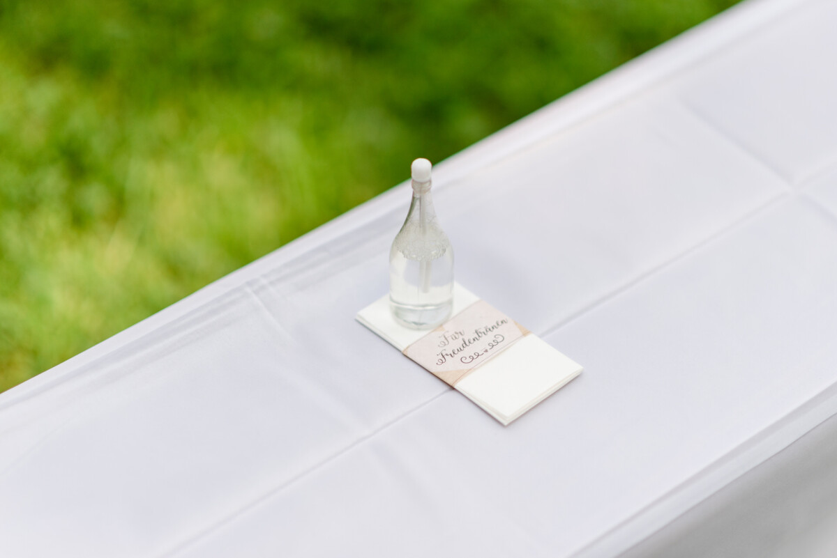 Ein mit einer weißen Tischdecke bedeckter Tisch, auf dem vor einem unscharfen grünen Hintergrund eine kleine Flasche Wasser und ein Umschlag liegen.