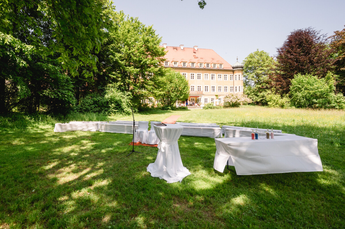 Veranstaltungsaufbau im Freien mit weiß gedeckten Tischen, einem Mikrofonständer und einem roten Teppich auf einer Rasenfläche. Im Hintergrund ist ein großes Gebäude mit rotem Dach zu sehen, umgeben von Bäumen.