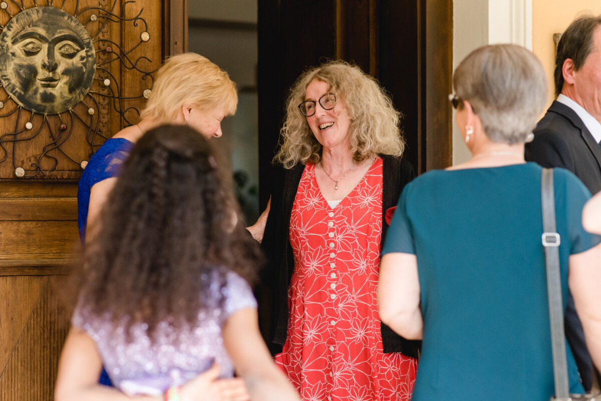 Eine Frau in einem roten Kleid und mit Brille lächelt, während sie sich in der Nähe einer Tür mit einer Gruppe von Menschen unterhält.