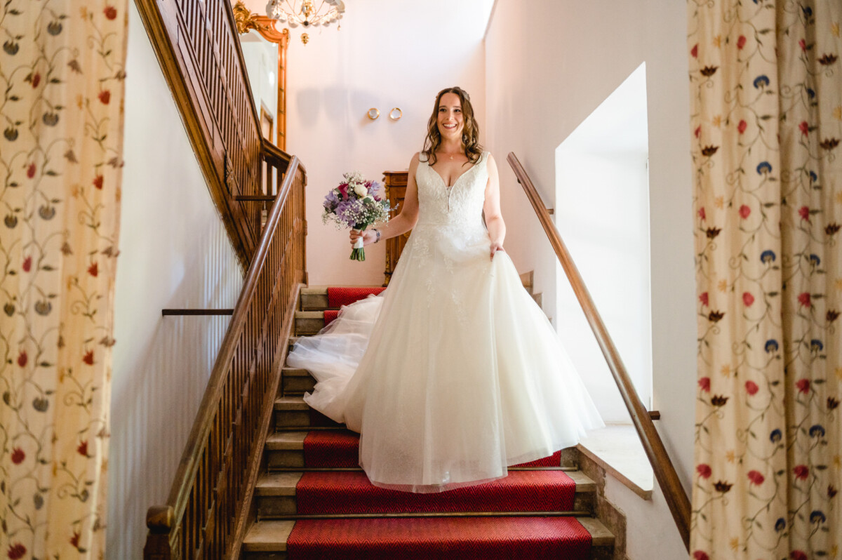 Eine Braut kommt im Brautkleid eine Treppe herunter und hält einen Blumenstrauß in der Hand.
