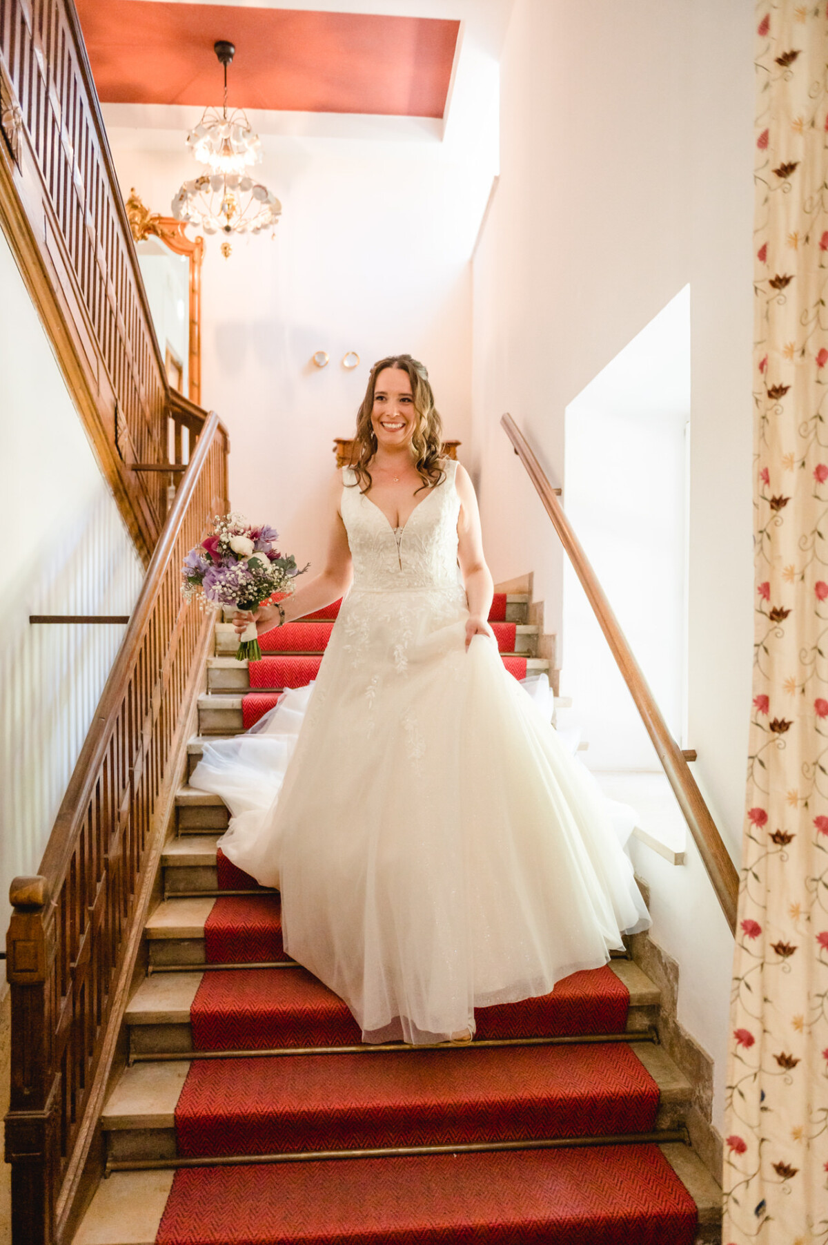 Eine Braut im weißen Brautkleid kommt lächelnd und mit einem Blumenstrauß in der Hand eine Treppe herunter.