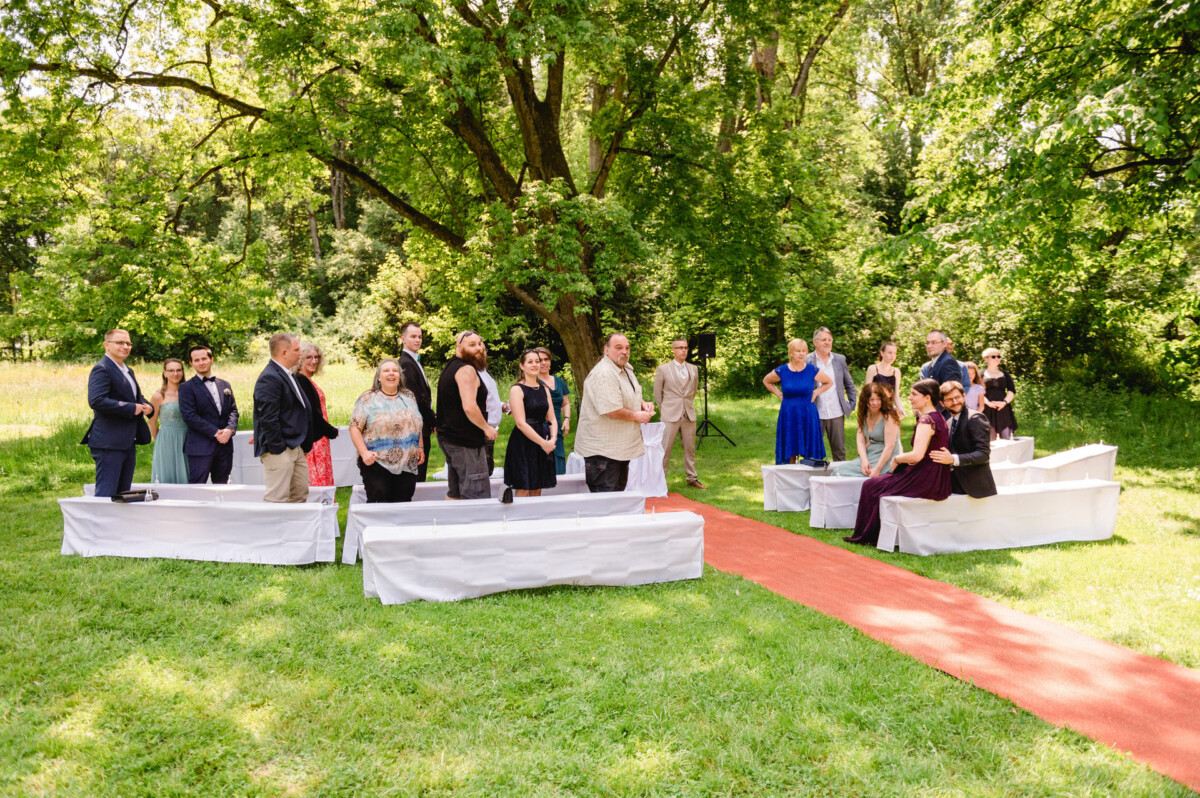 Eine kleine Gruppe von Menschen steht und sitzt auf weißen Bänken bei einem Treffen im Freien auf einer Rasenfläche mit einem roten Teppich, umgeben von Bäumen.