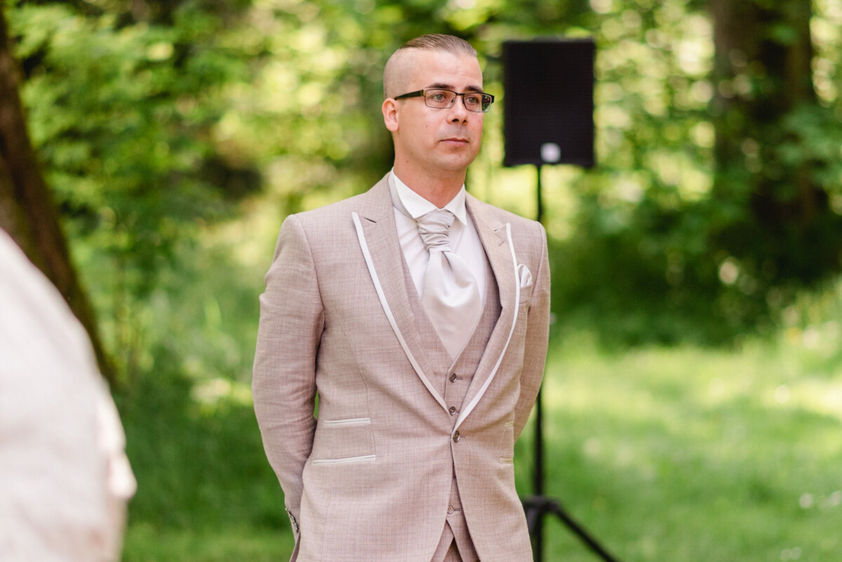 Ein Mann in einem hellbeigen Anzug steht im Freien, im Hintergrund sind Bäume und ein Lautsprecher zu sehen.