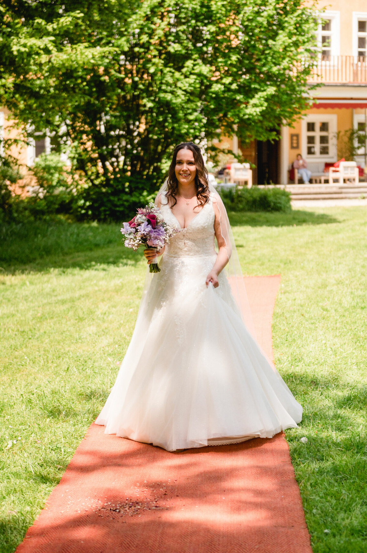 Eine Braut in einem weißen Kleid schreitet mit einem Blumenstrauß in der Hand über einen roten Teppich im Freien zum Altar entlang. Im Hintergrund sind Grünpflanzen und ein Gebäude zu sehen.