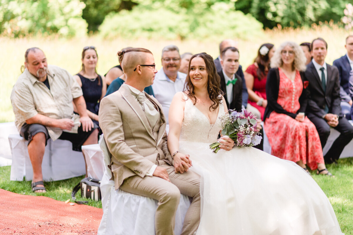 Braut und Bräutigam sitzen bei einer Hochzeitszeremonie im Freien Händchen haltend zusammen, umgeben von Gästen, die auf weißen Stühlen sitzen. Die Braut hält einen Blumenstrauß.