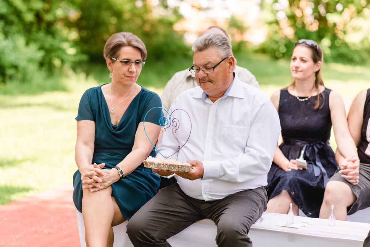 Ein Mann und eine Frau sitzen im Freien. Der Mann hält einen Kuchen mit Herzverzierung. Hinter ihnen sitzen zwei Personen.